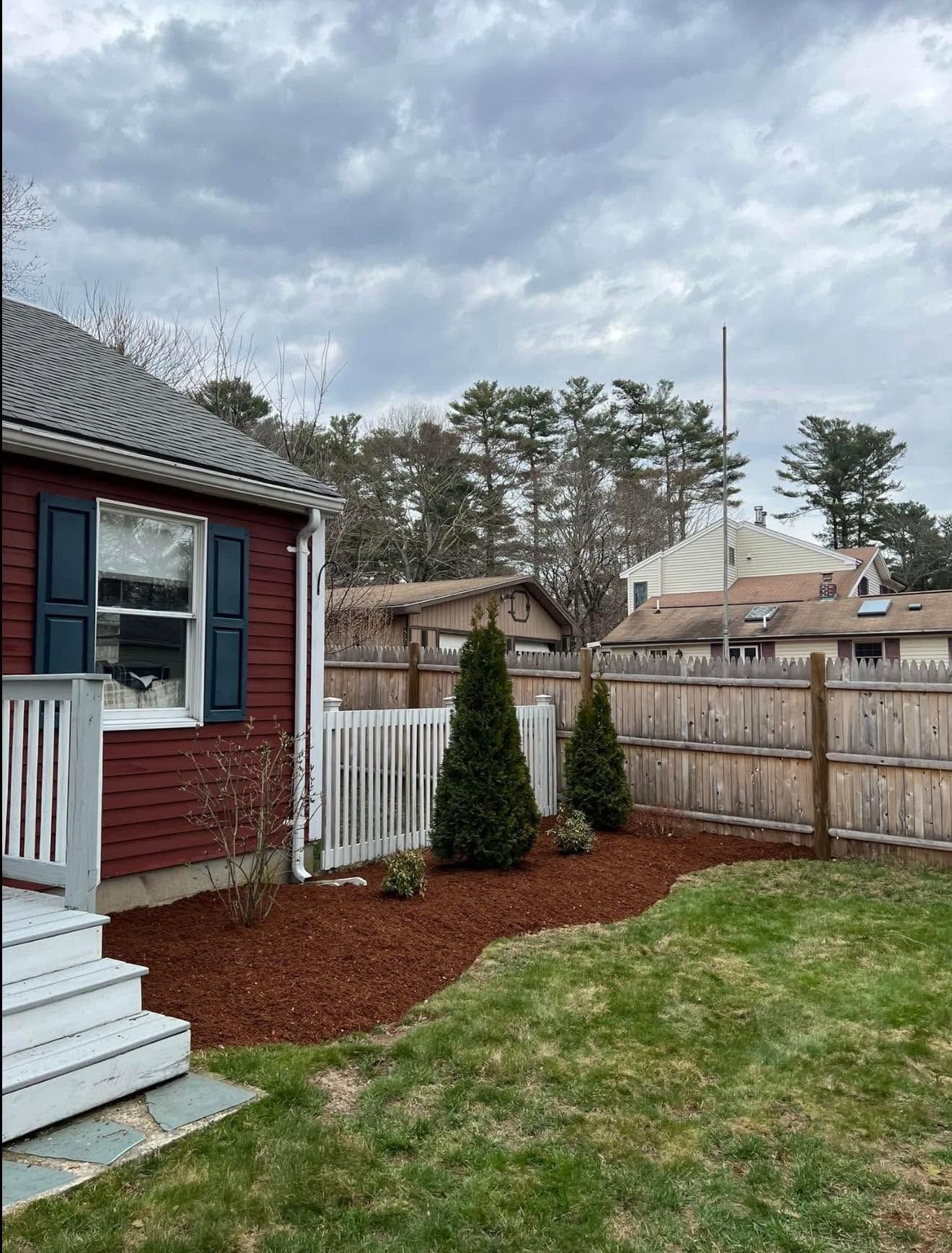 Backyard beside red house with white porch, small evergreens, grass, and wooden fence under cloudy sky