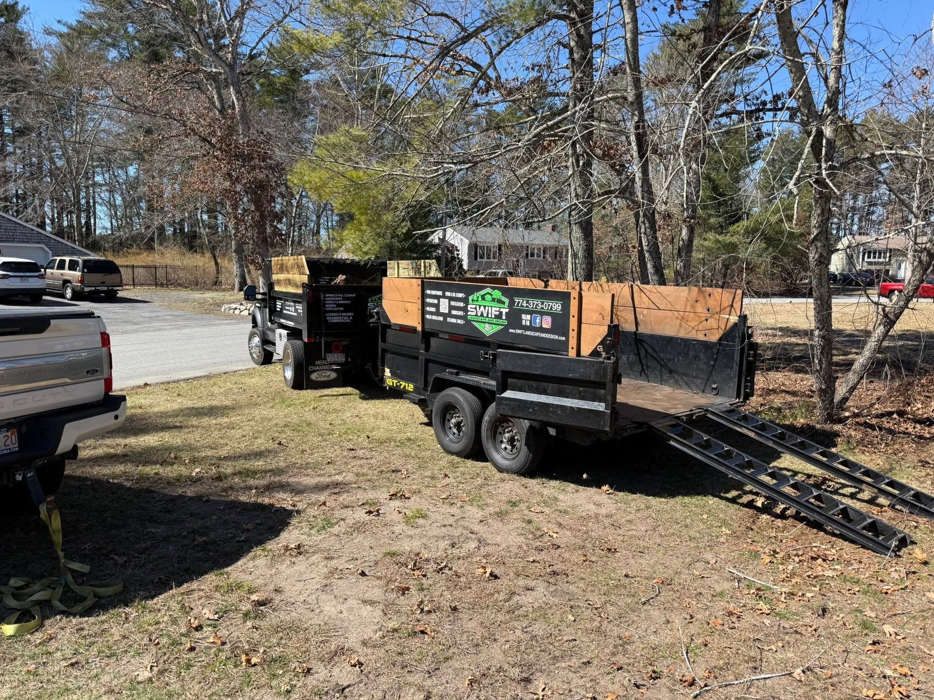 Black utility trailer with ramp parked in a yard beside a white pickup truck on a sunny day.