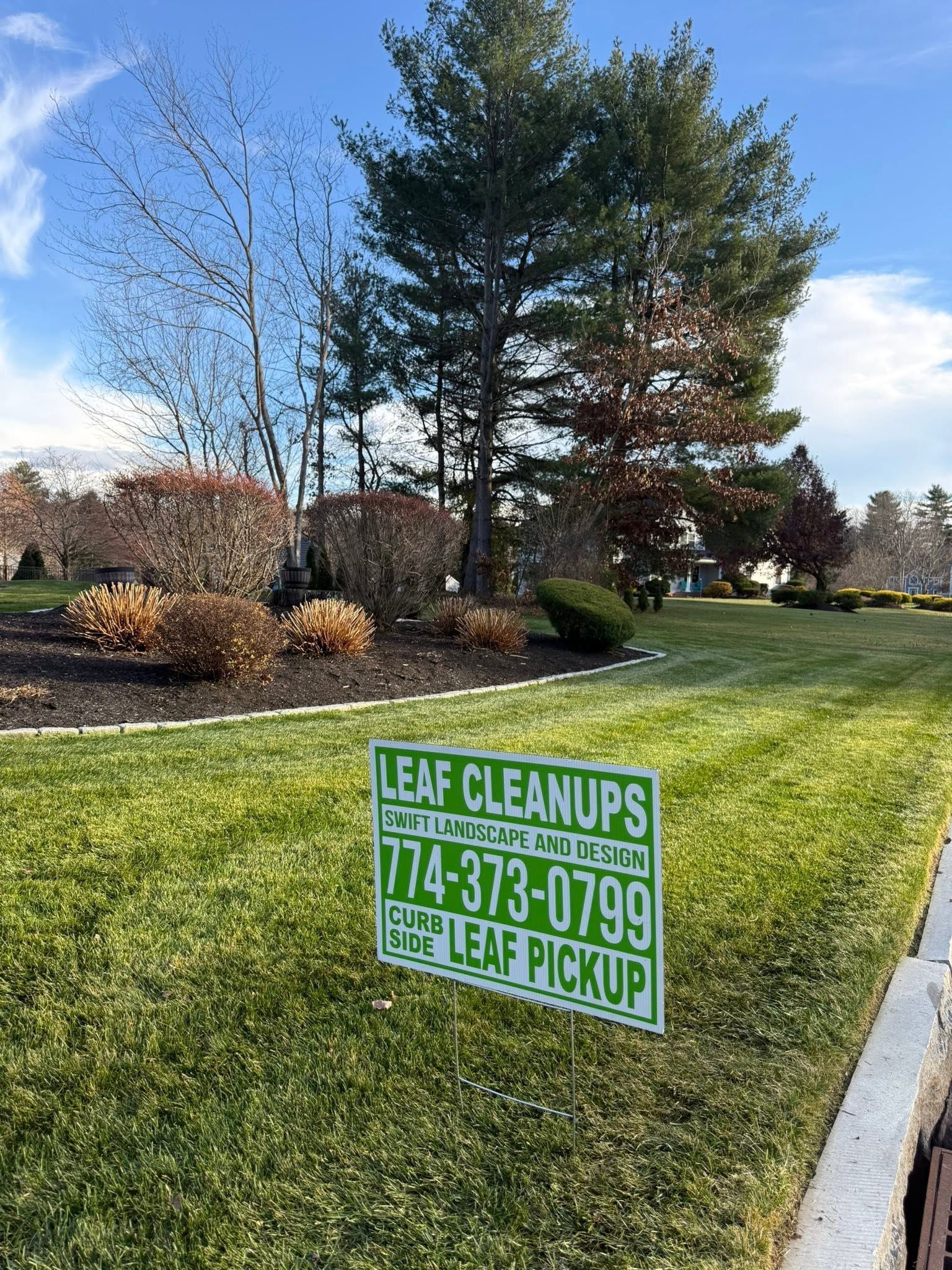 Leaf Cleanups yard sign on a grassy roadside beside trees under a blue sky.