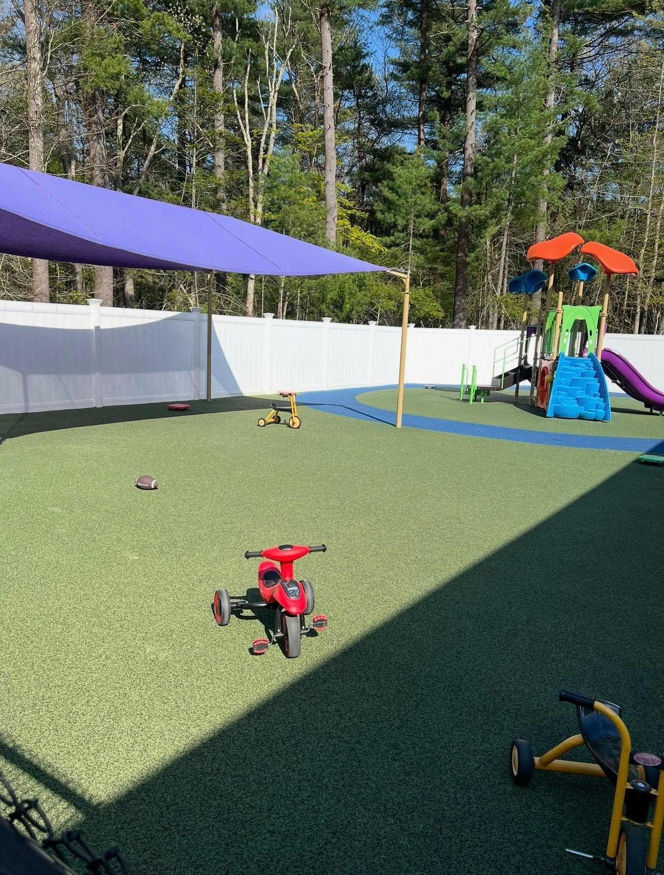Children’s playground with green turf, slides, and climbing equipment beside a white fence and trees