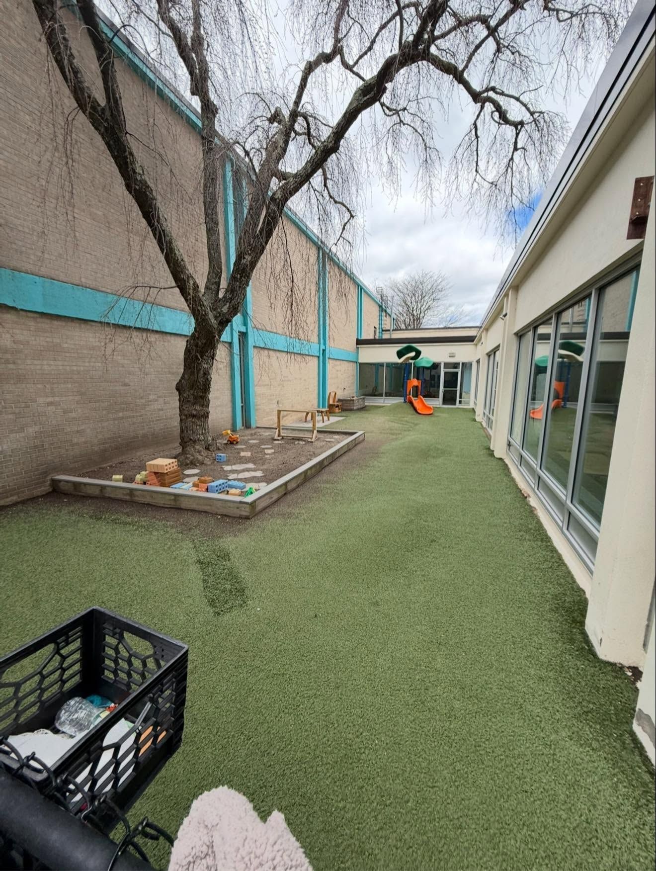 Narrow outdoor courtyard with green turf, a tree, raised bed, and a child near a swing set.