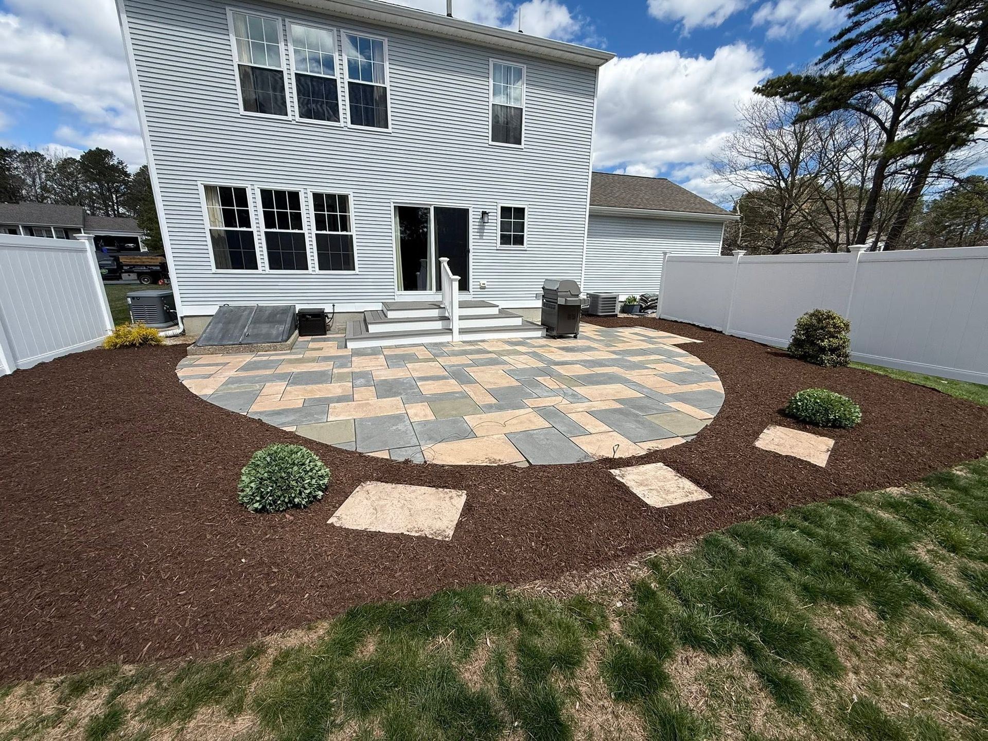 Backyard patio with stone seating area, mulch beds, and white house exterior under a blue sky