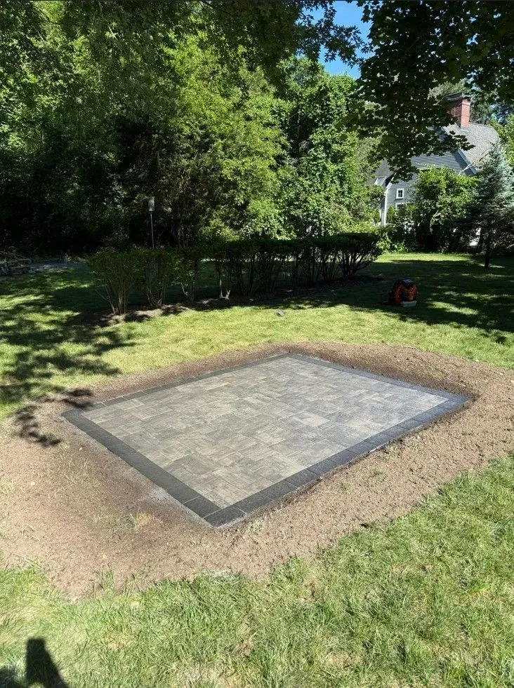 Rectangular concrete pad bordered in dark trim on a grassy yard, with trees and a house in the background
