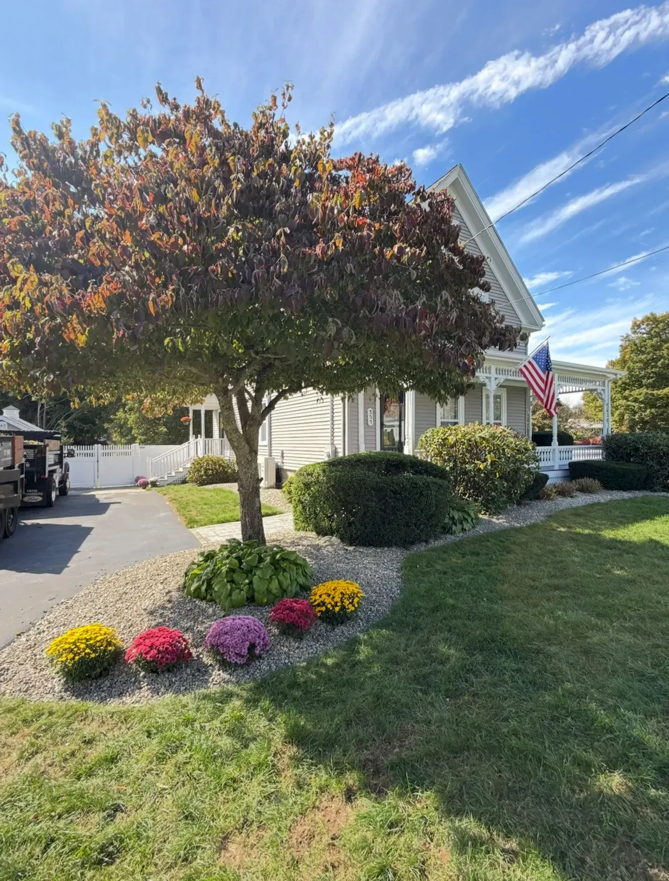 House with landscaped front yard, colorful flower bed, trimmed bushes, and an American flag under a blue sky