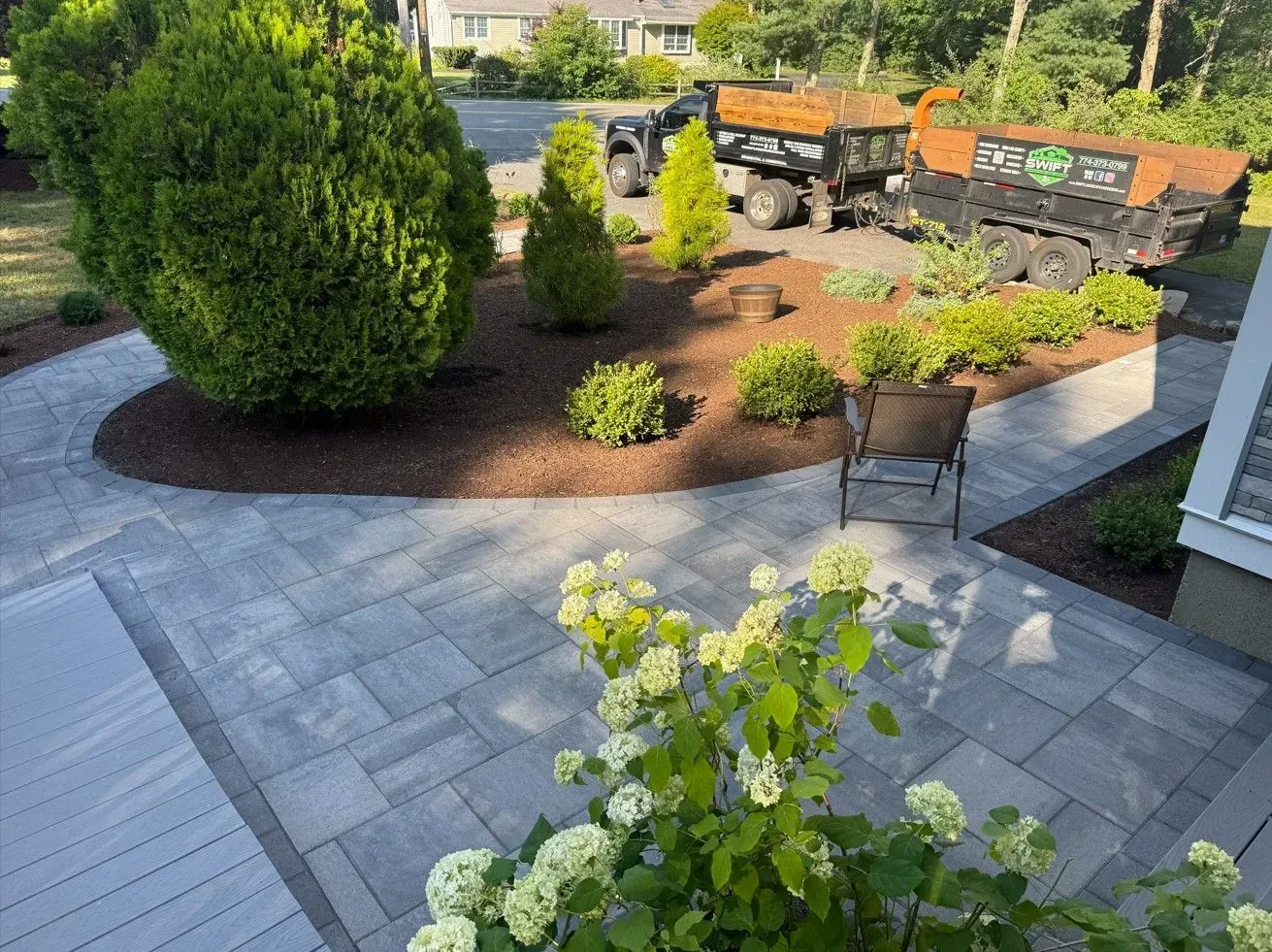Landscaped courtyard with gray pavers, mulch beds, shrubs, and parked trucks in the background