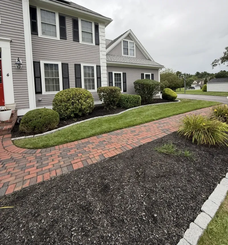 Gray house with black shutters, red brick walkway, and trimmed green shrubs in a front yard