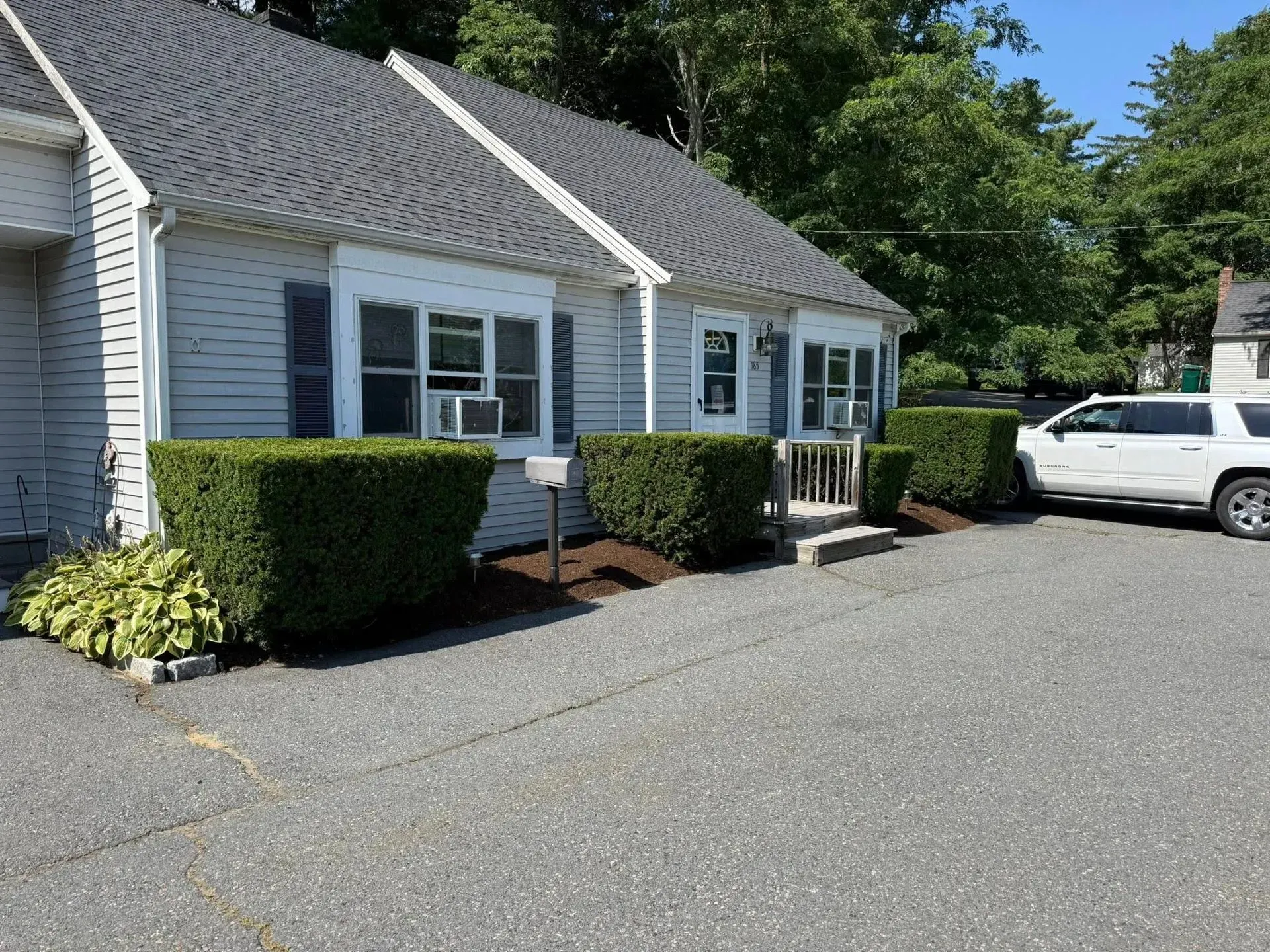 White house with blue shutters, trimmed hedges, and a parked SUV in a paved lot