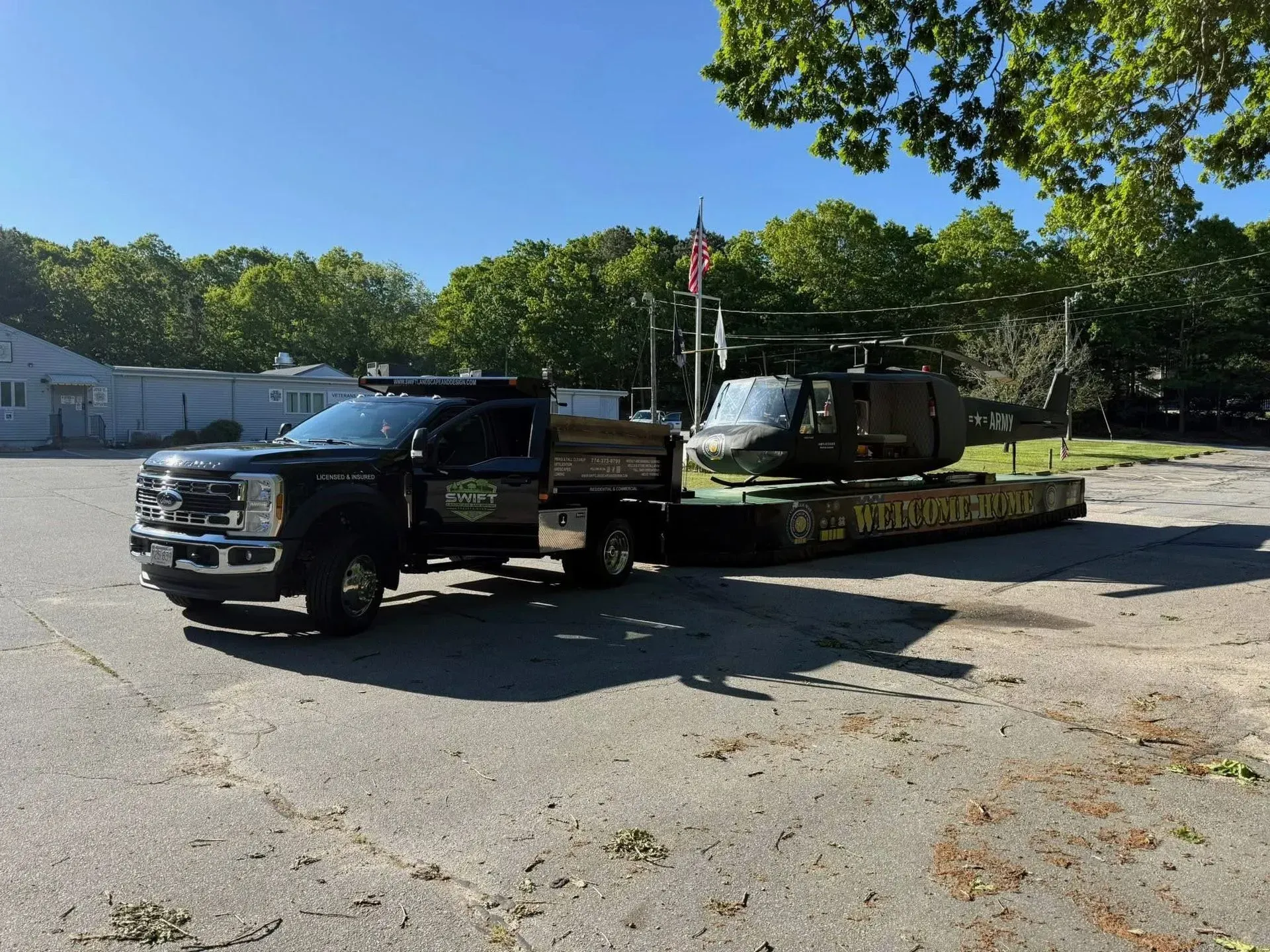 Two people standing beside a black helicopter and trailer in a sunny parking lot