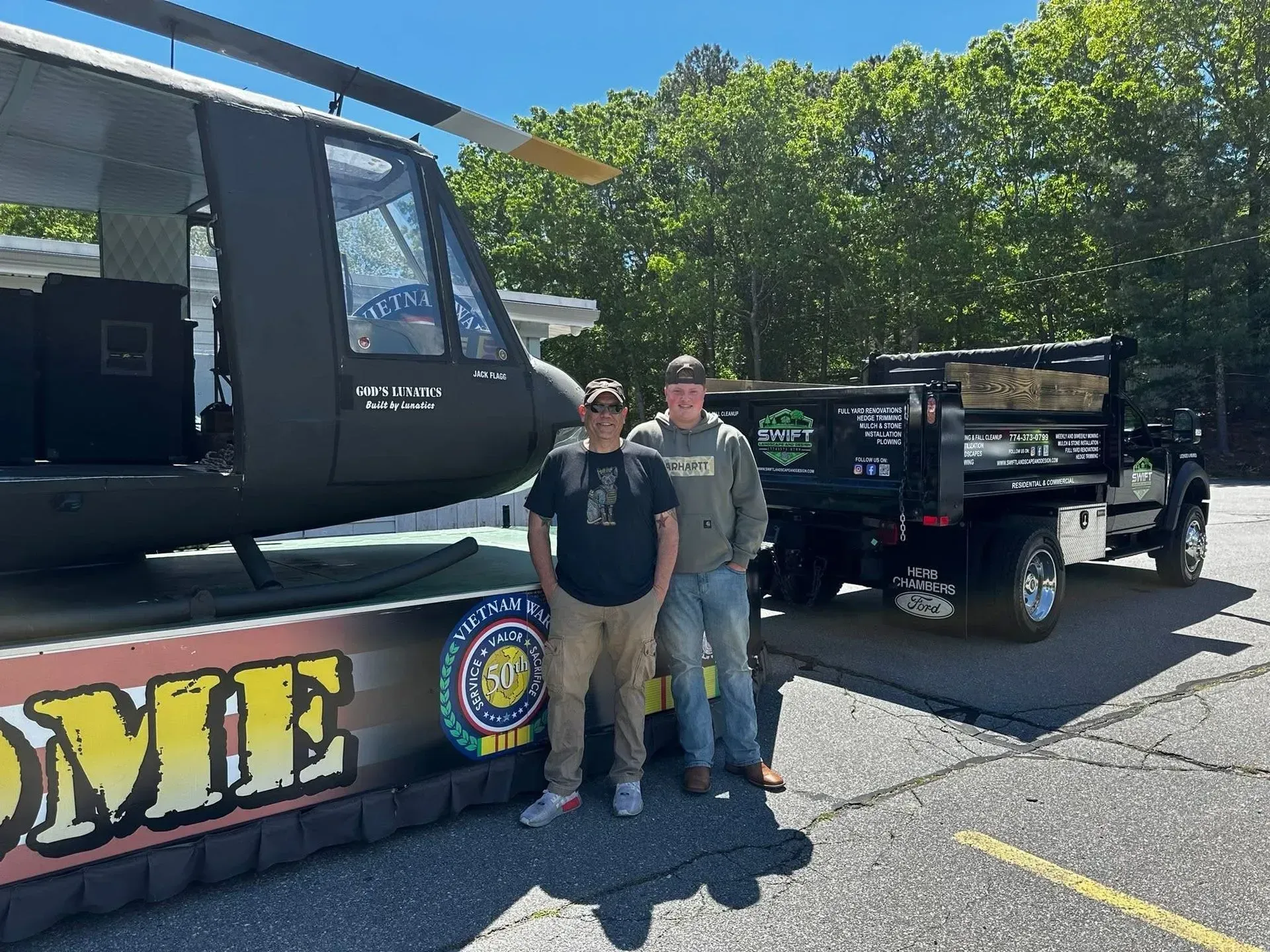 Two people standing beside a black helicopter and trailer in a sunny parking lot