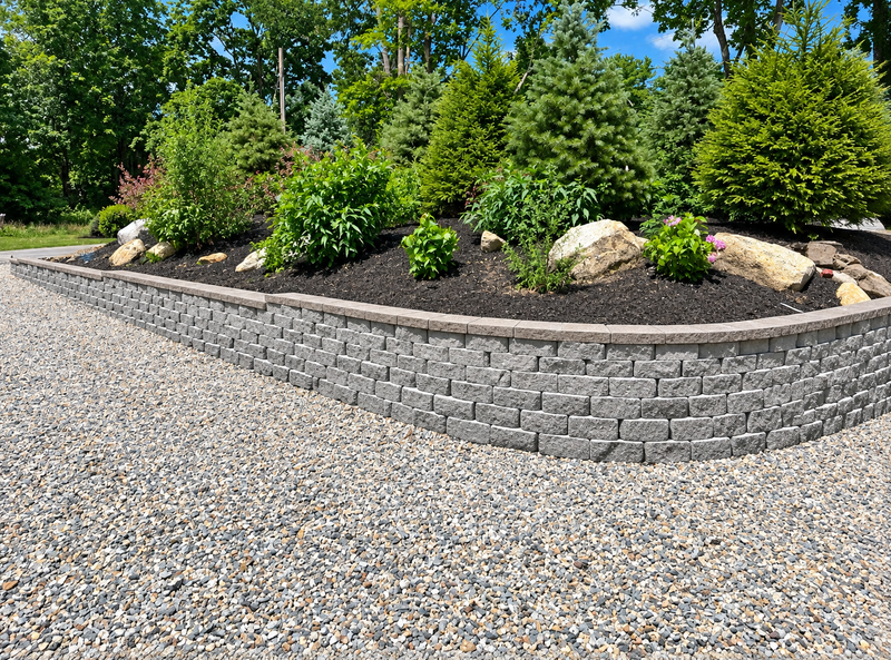 Landscaped retaining wall with shrubs and rocks beside a gravel driveway under blue sky