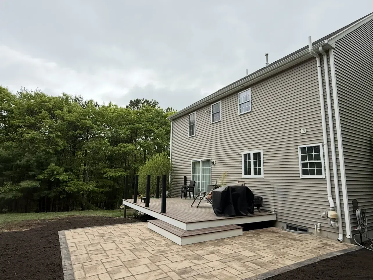 Back patio with paver steps, black railings, grill cover, and beige siding beside a wooded yard