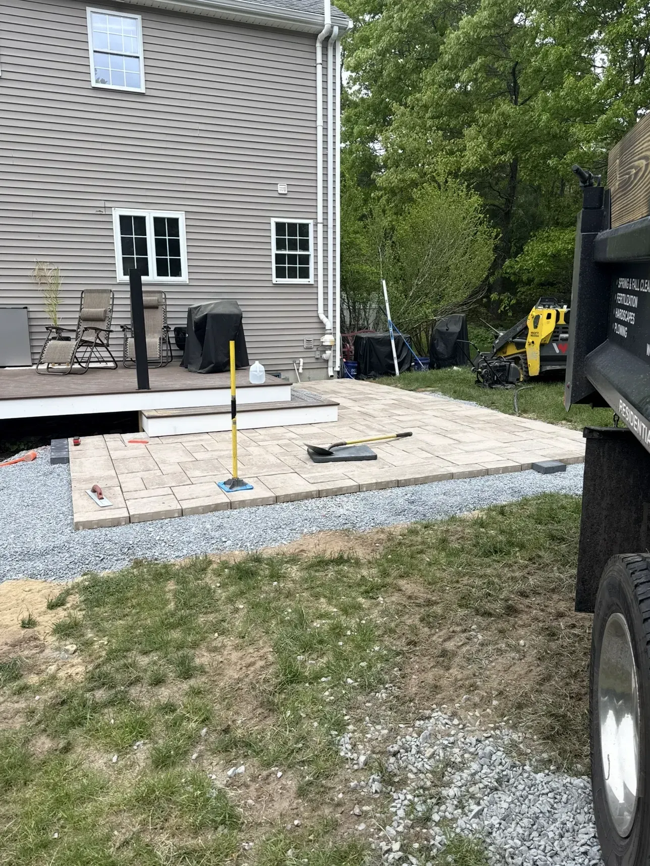 Backyard patio under construction beside a house, with pavers, gravel, and tools nearby.