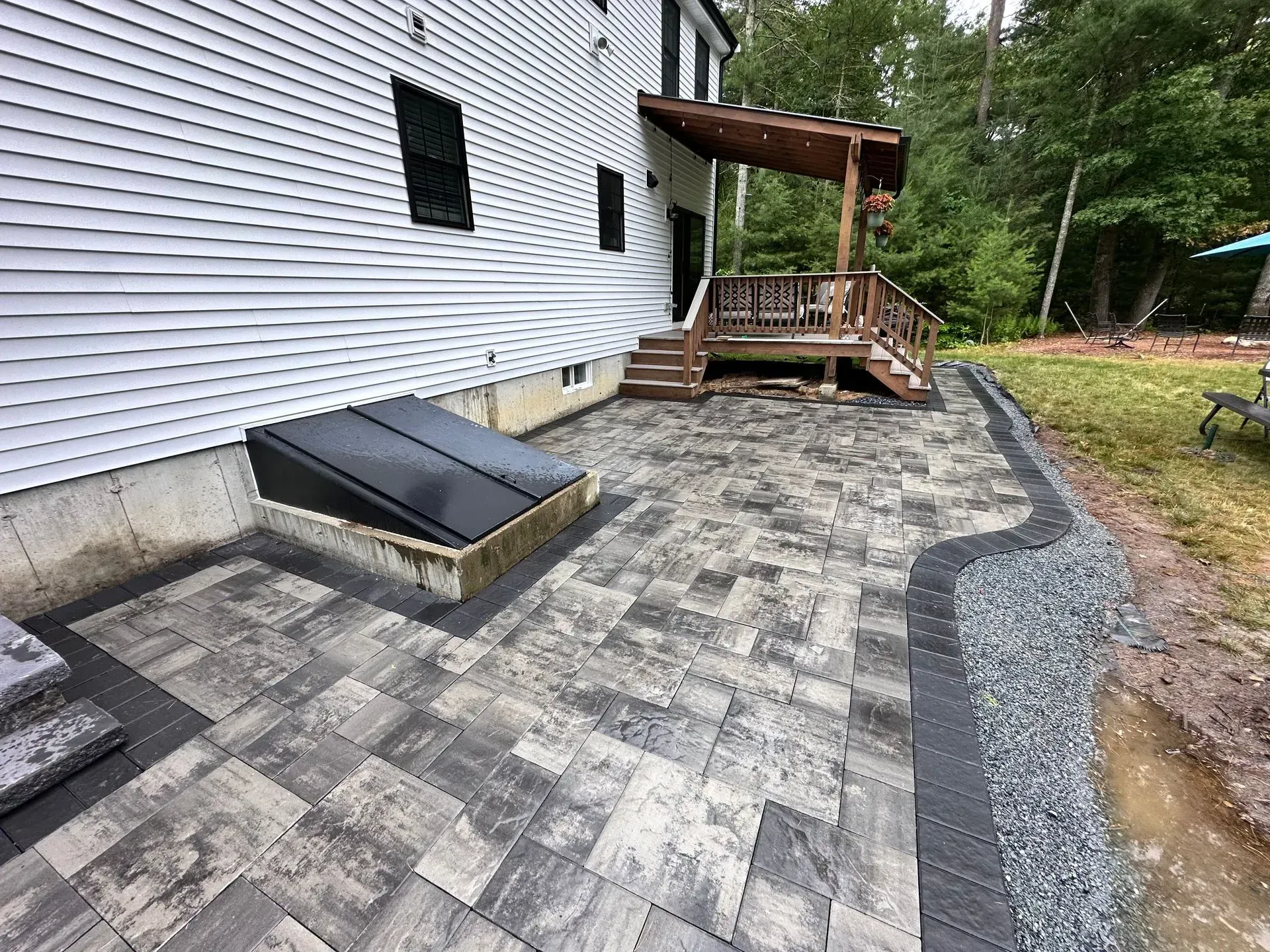 Patio with gray pavers beside white house, black basement window well, and small wooden deck with stairs