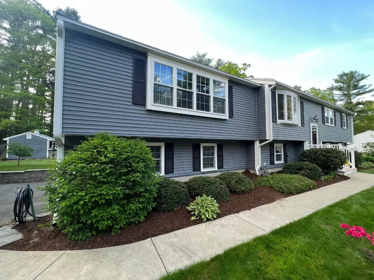 Gray split-level house with white-trim windows, shrubs, sidewalk, and a small lawn by the street
