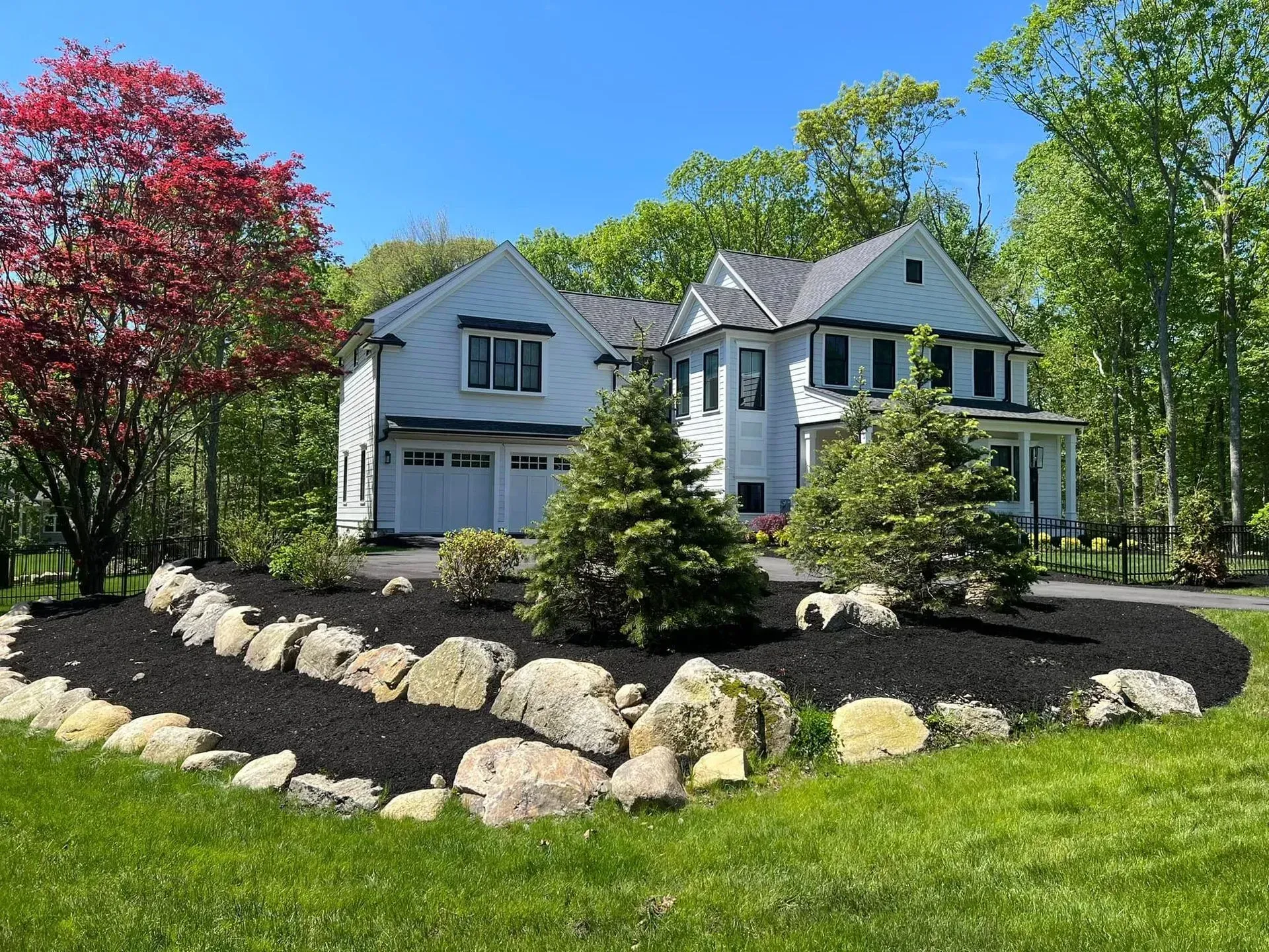 White house with gray roof in a sunny yard, framed by trees and a curved rock garden.
