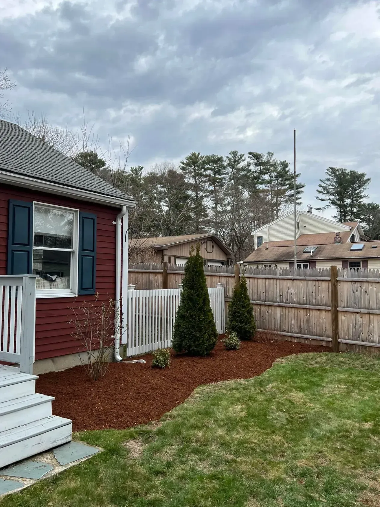 Backyard beside a red house with white fence, two shrubs, and cloudy sky