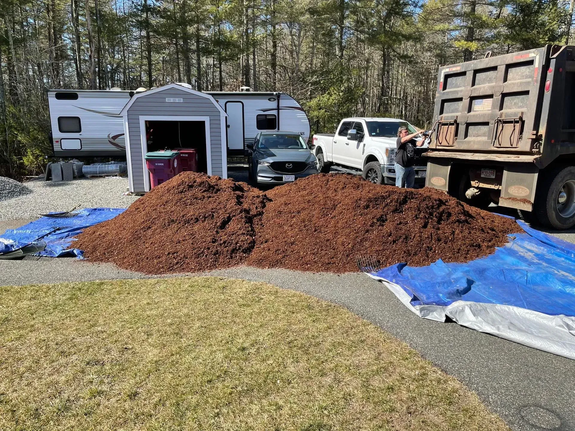 Mulch pile on blue tarp in driveway, with RV, van, and dump truck in the background