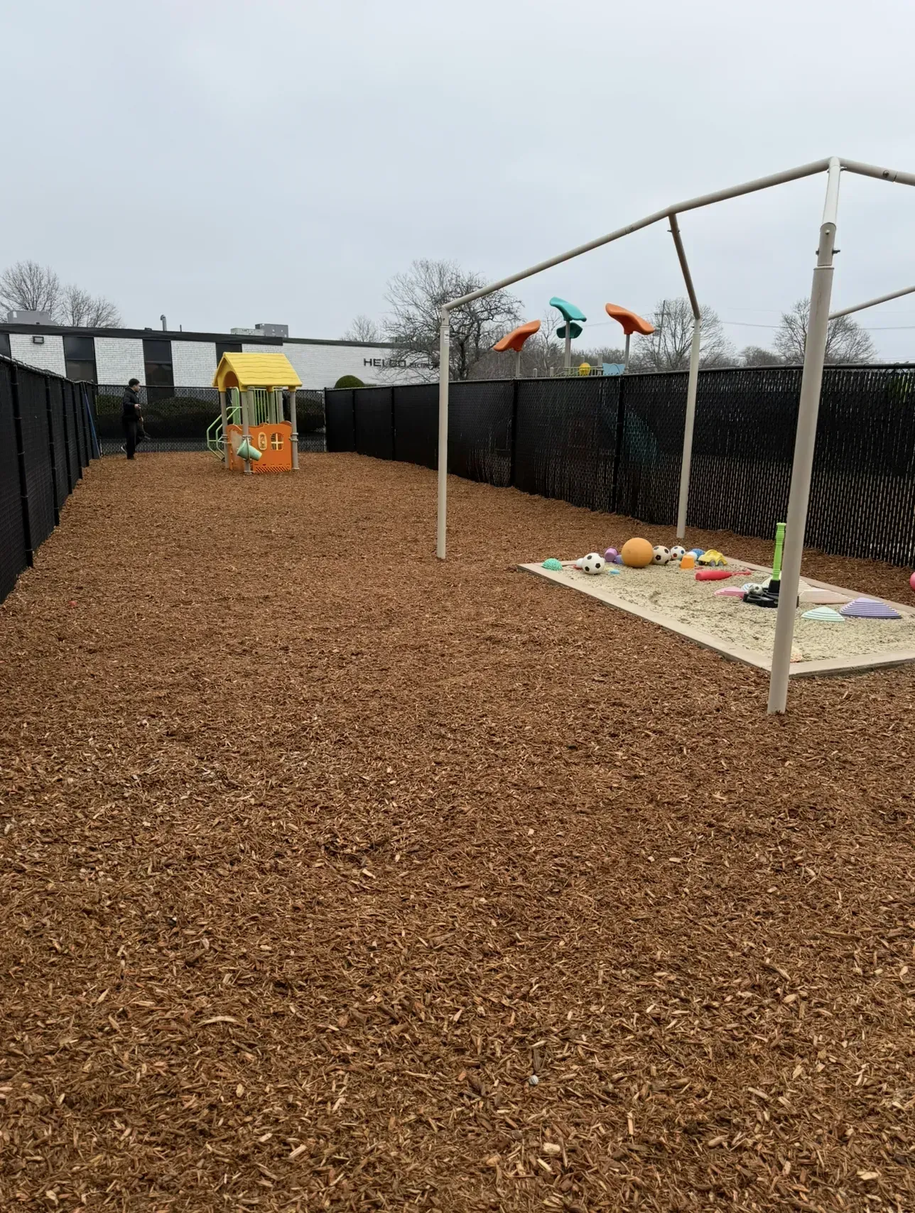Outdoor play yard with wood chips, a small playhouse, and a canopy-covered mat area with toys under a cloudy sky