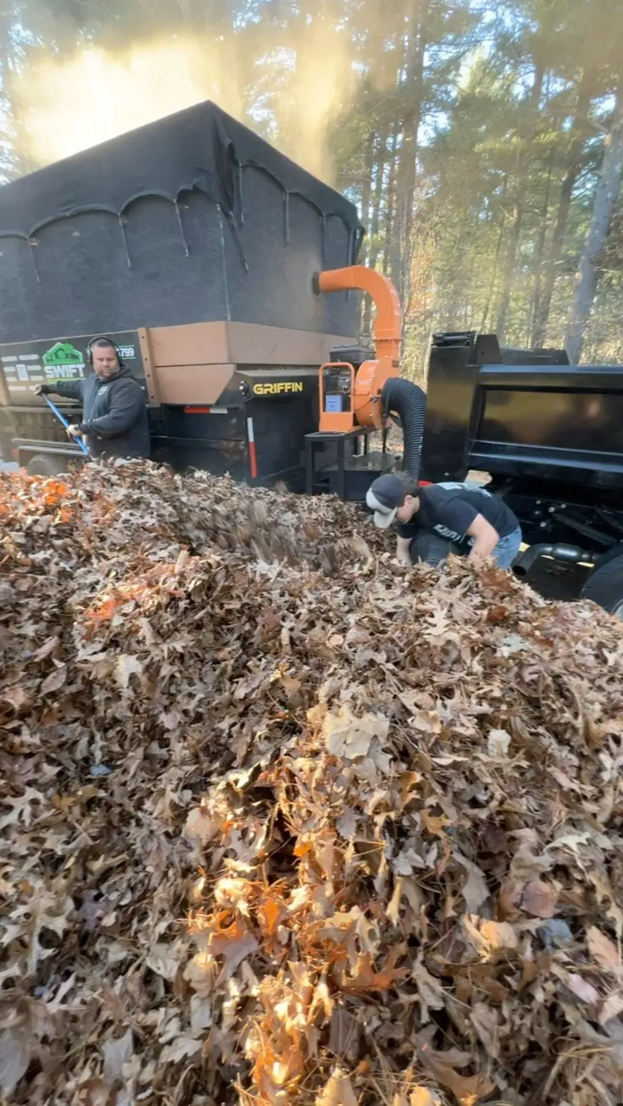 Workers using a leaf vacuum to clear a large pile of autumn leaves near a truck in a wooded area