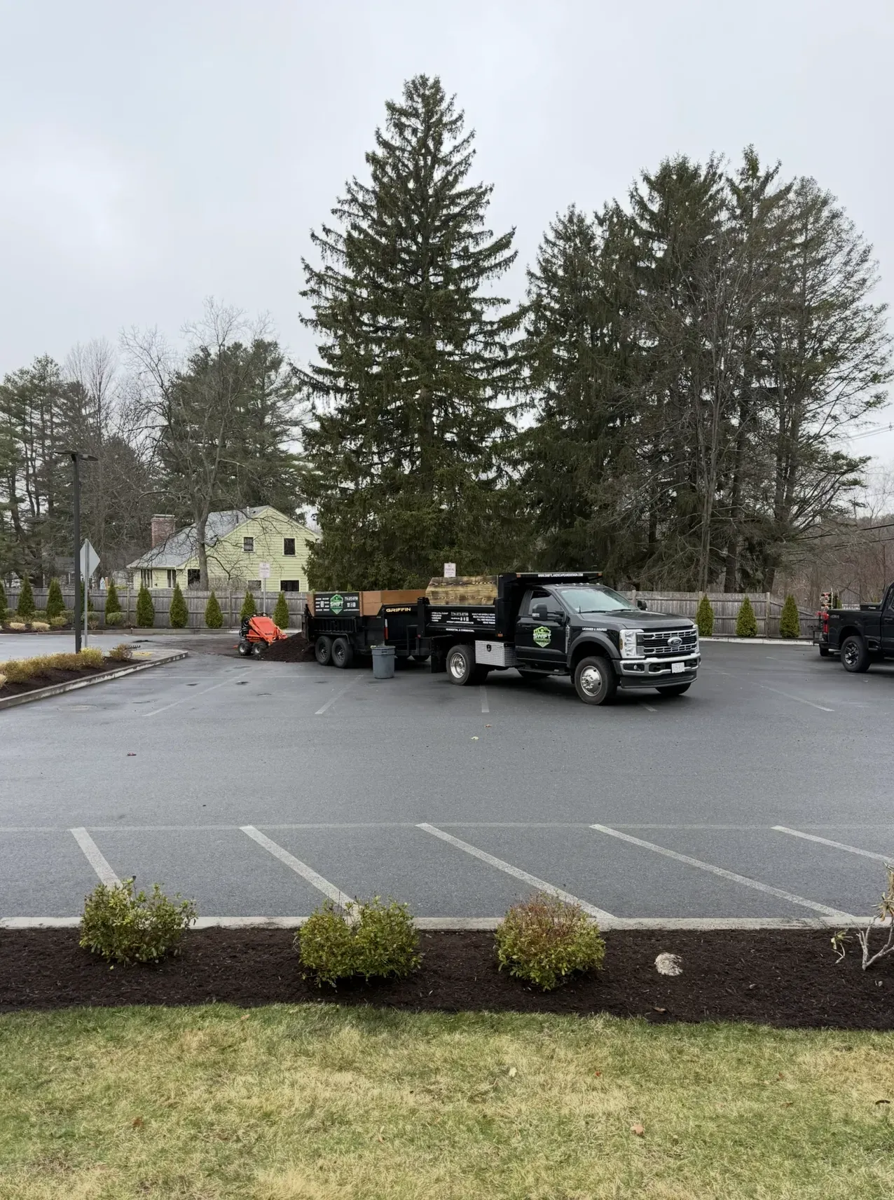 Truck with flatbed trailer in a wet parking lot, with workers and trees in the background