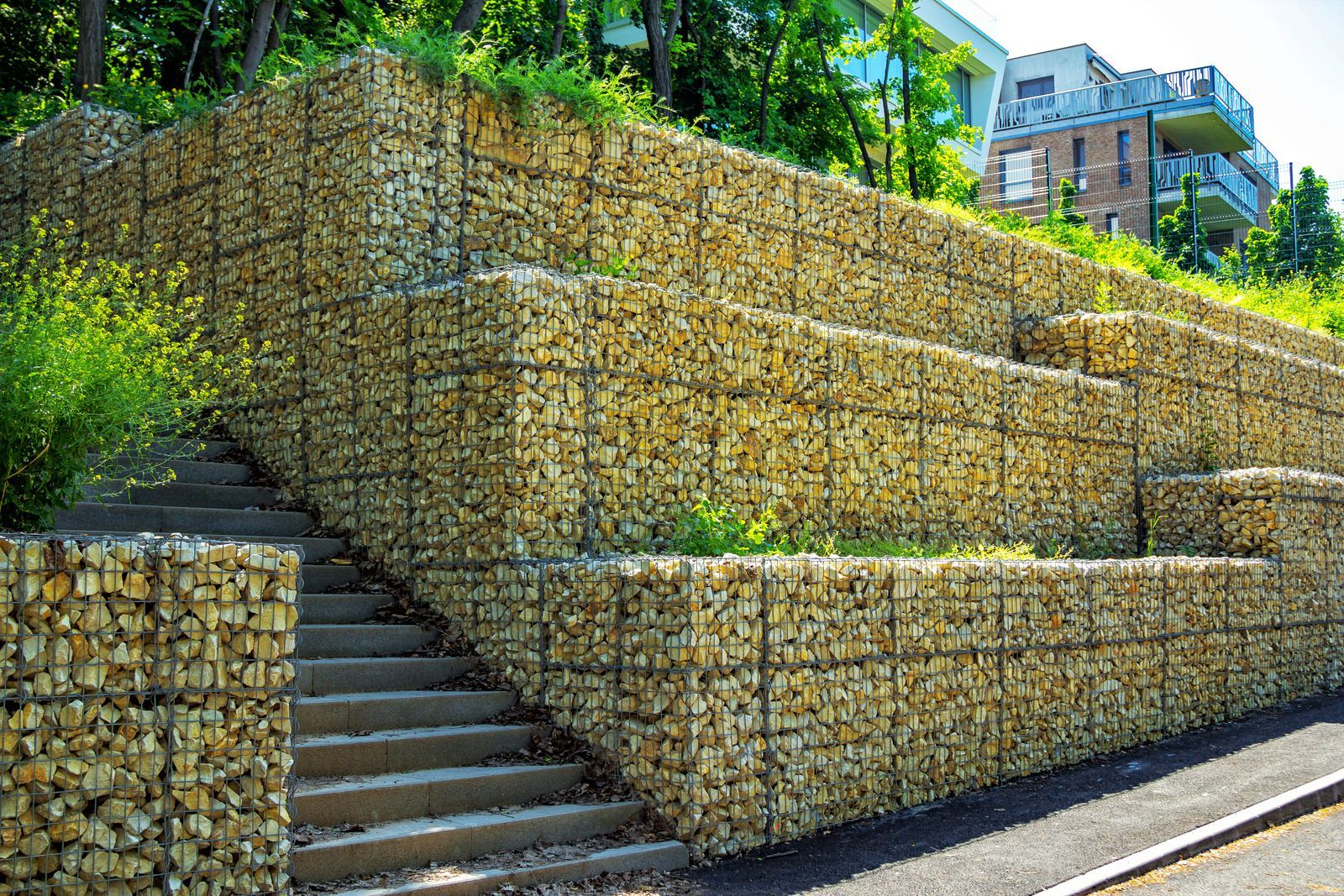 Gabion retaining walls and stairs beside a road, with greenery and buildings above.