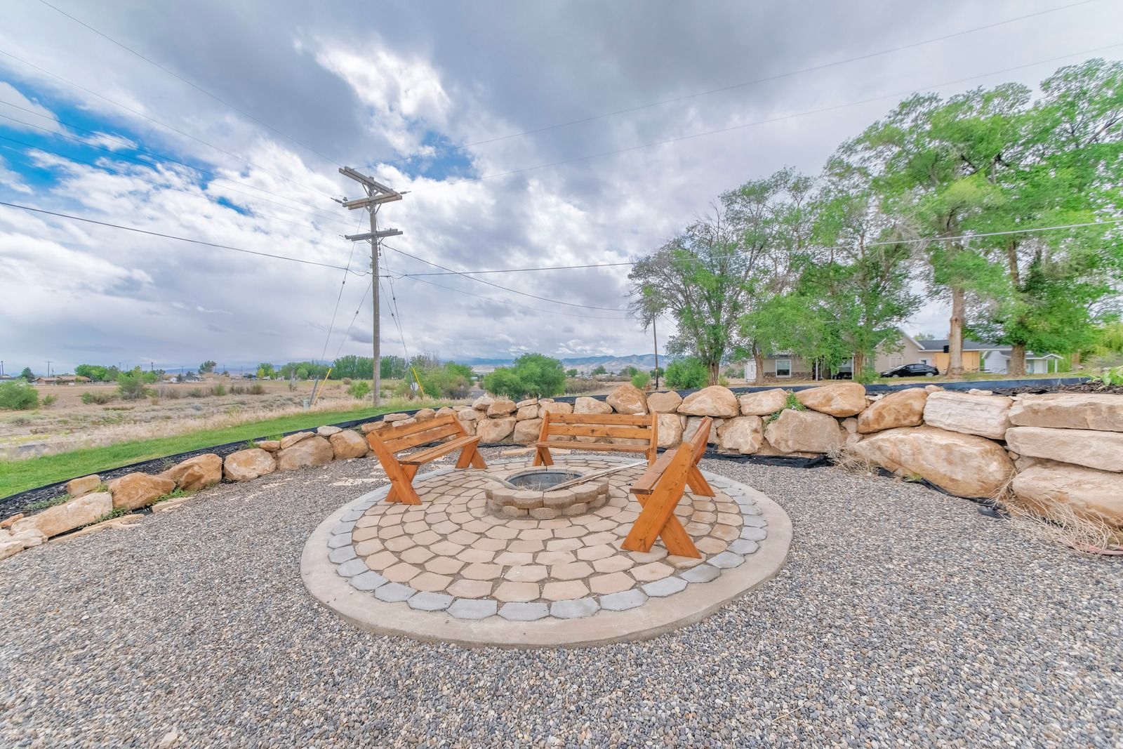 Outdoor stone fire pit seating area with wooden chairs, gravel ground, and open rural landscape under cloudy sky