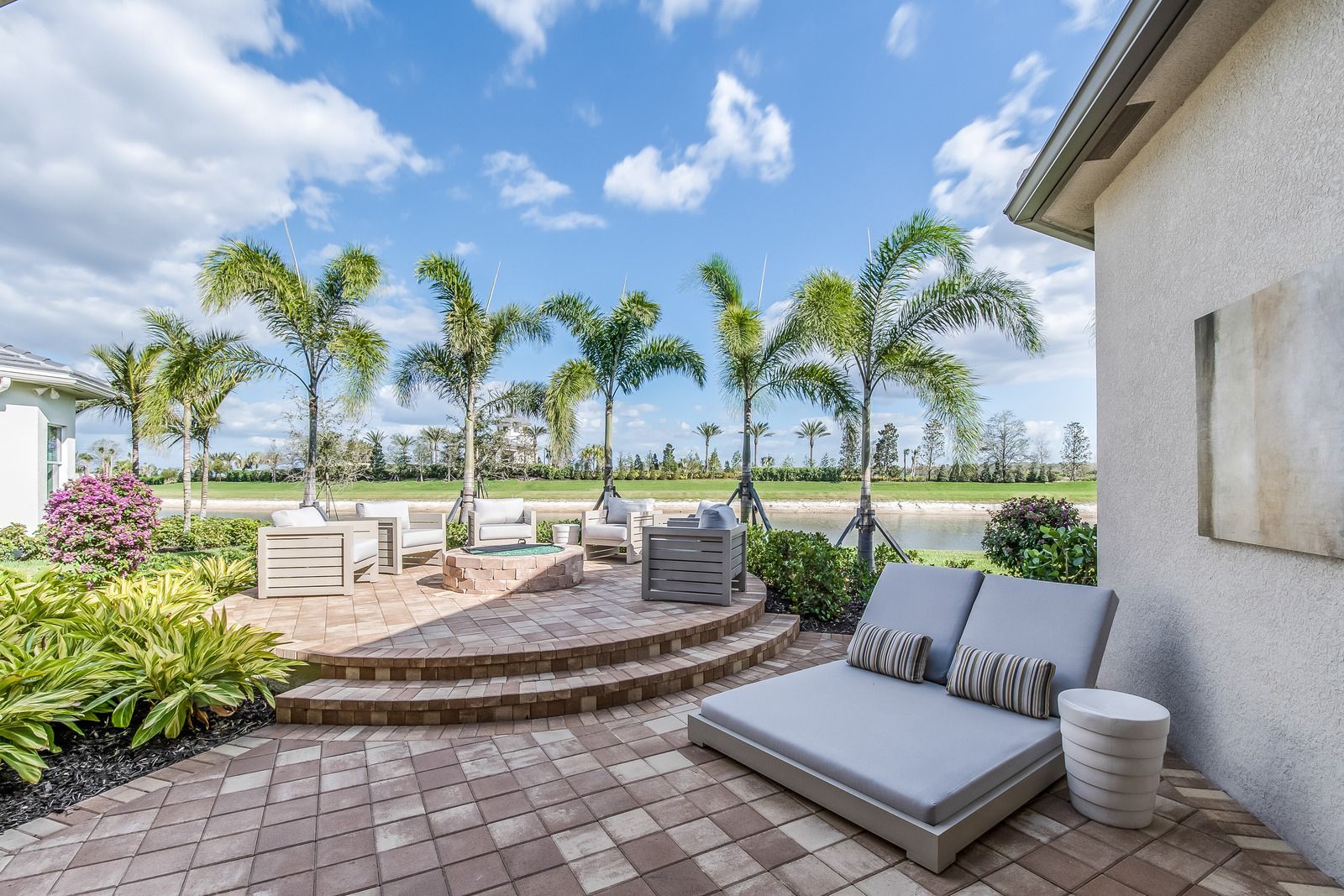 Backyard patio with gray lounge chairs, potted plants, palm trees, and a bright ocean view