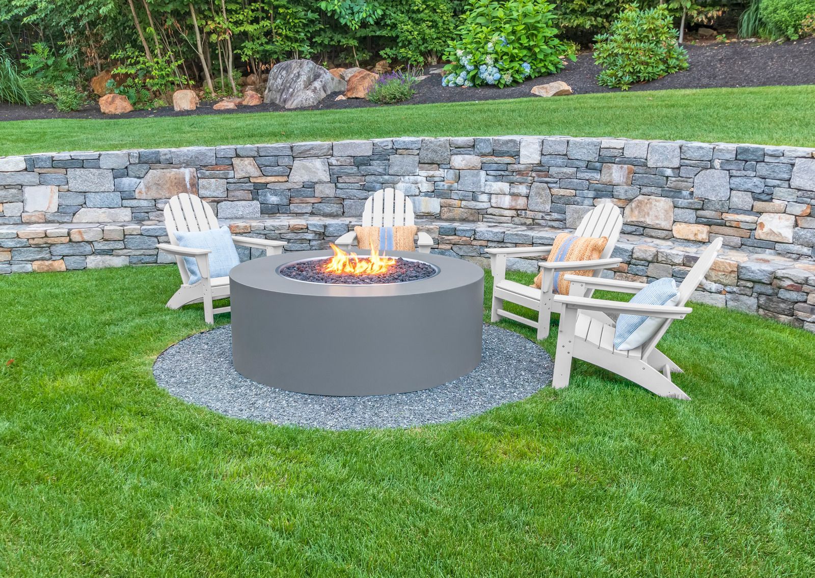Backyard fire pit surrounded by white chairs on green lawn beside a stone retaining wall