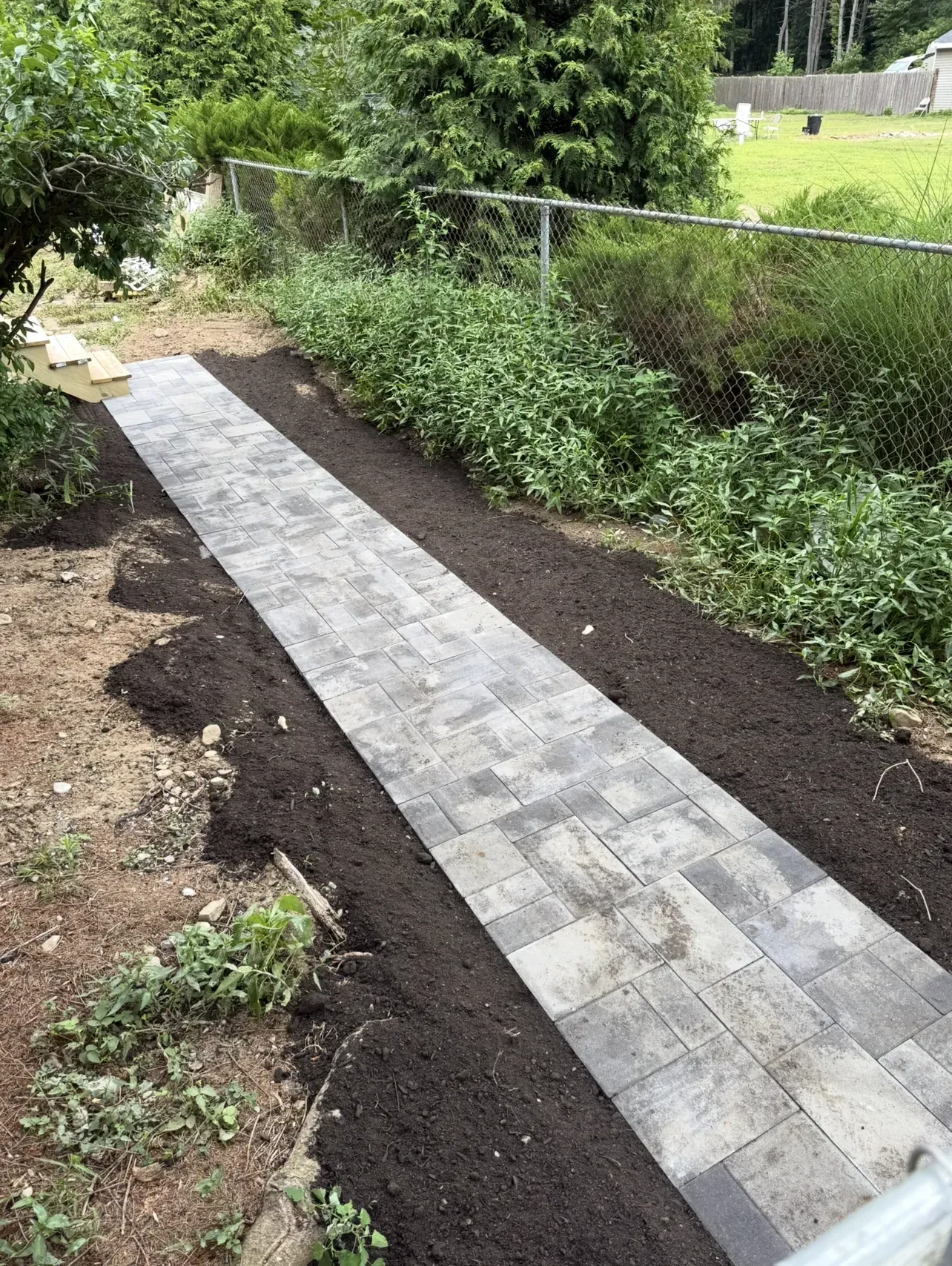 A narrow paved garden path beside dark soil beds and green shrubs, with a fence along the right side.