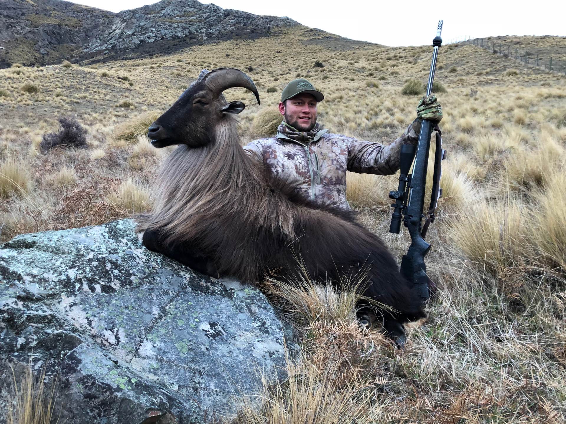 Man in camouflage gear poses with a large Himalayan tahr he has apparently hunted, rifle in hand, in a grassy, mountainous landscape.