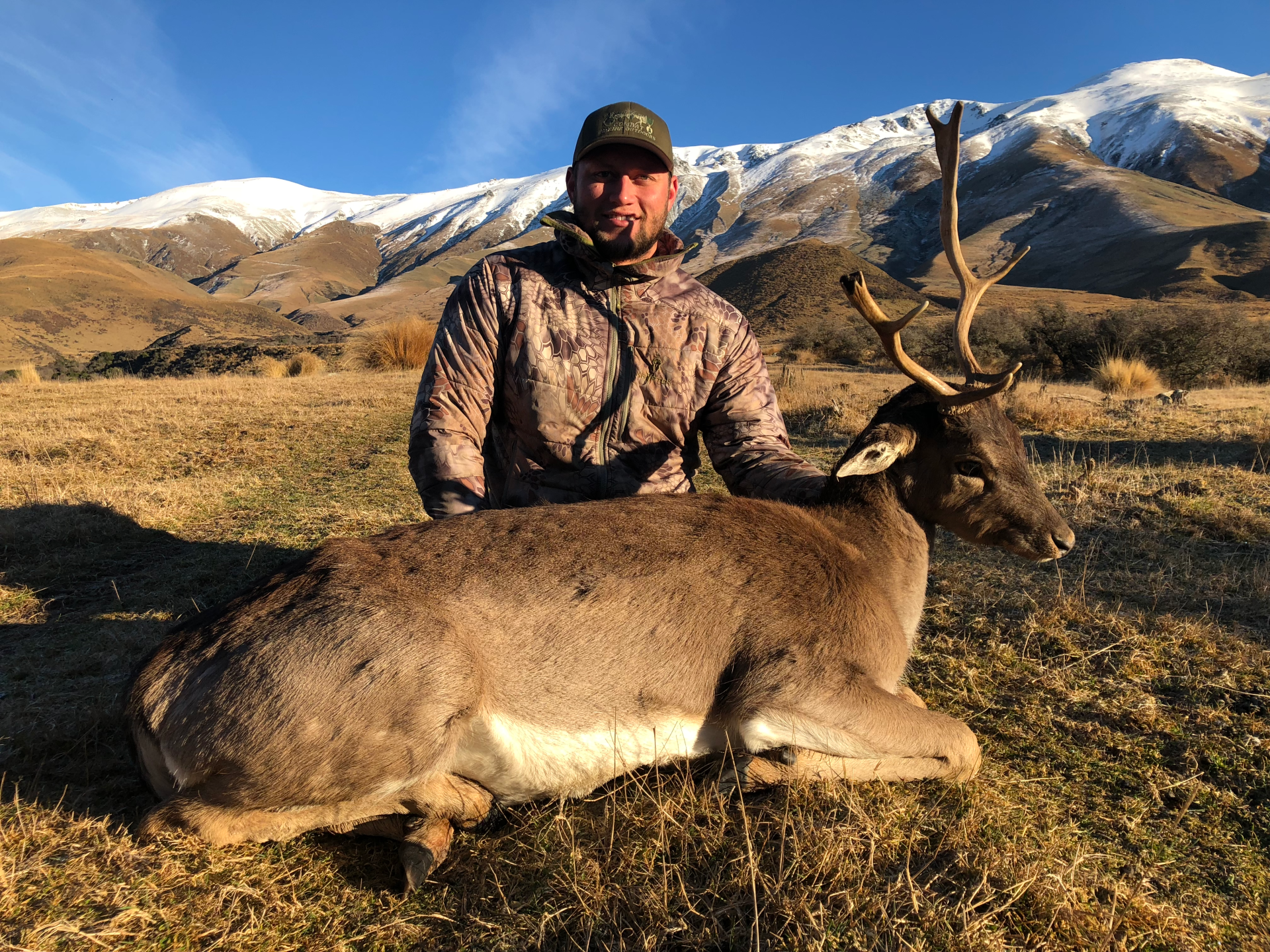 Man in camouflage with a harvested deer, antlers prominent, in a grassy field with snow-capped mountains in the background.
