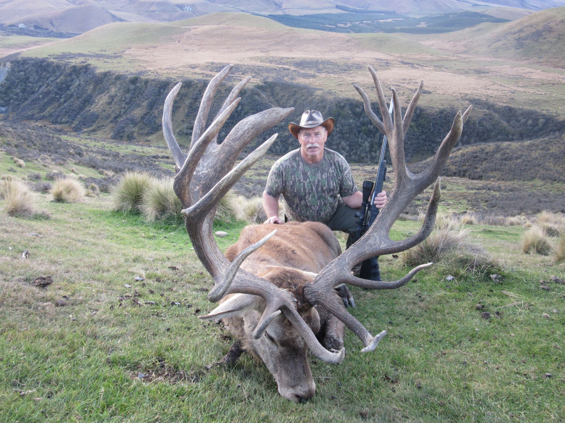 Hunter kneels beside a large dead red deer with enormous antlers, holding a rifle in a grassy hillside setting.