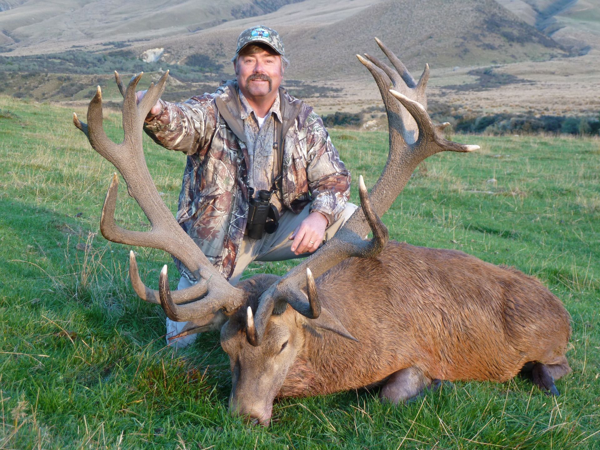 Man kneels beside a large deer with impressive antlers, in a grassy field. The hunter wears camouflage.