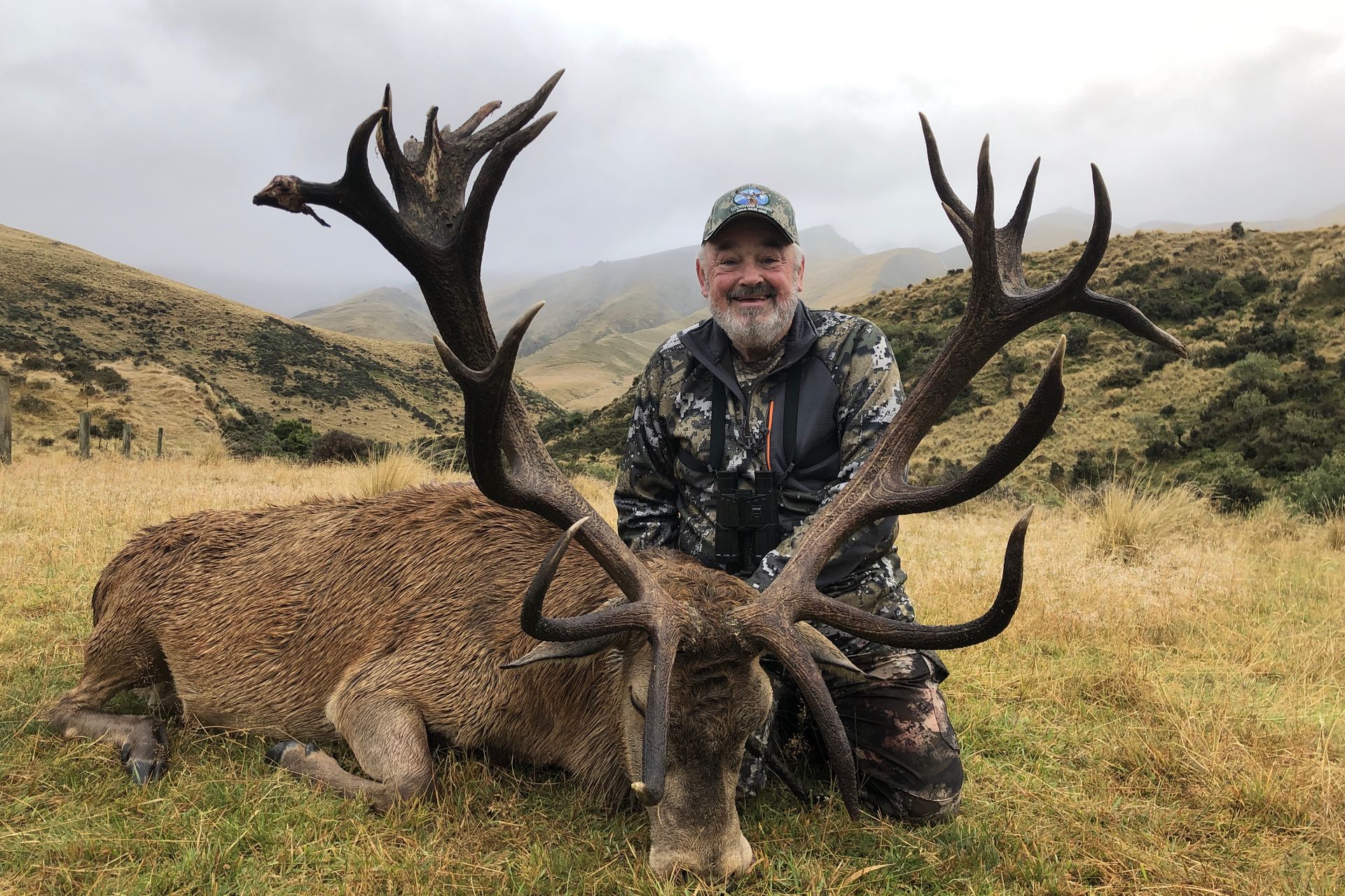 Man kneeling next to a large, dead red deer with impressive antlers, in a grassy mountain setting.
