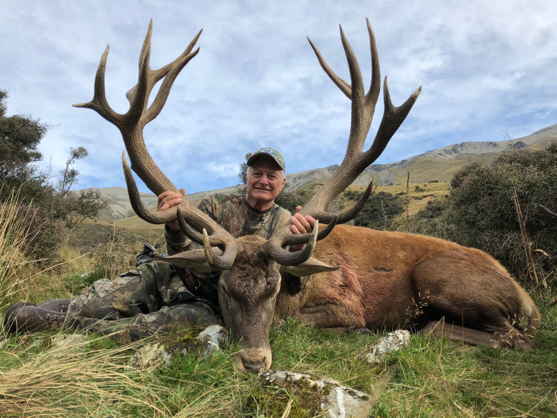 Hunter in camouflage holding large antlers of a red deer he has shot, in a grassy field with mountains in the background.