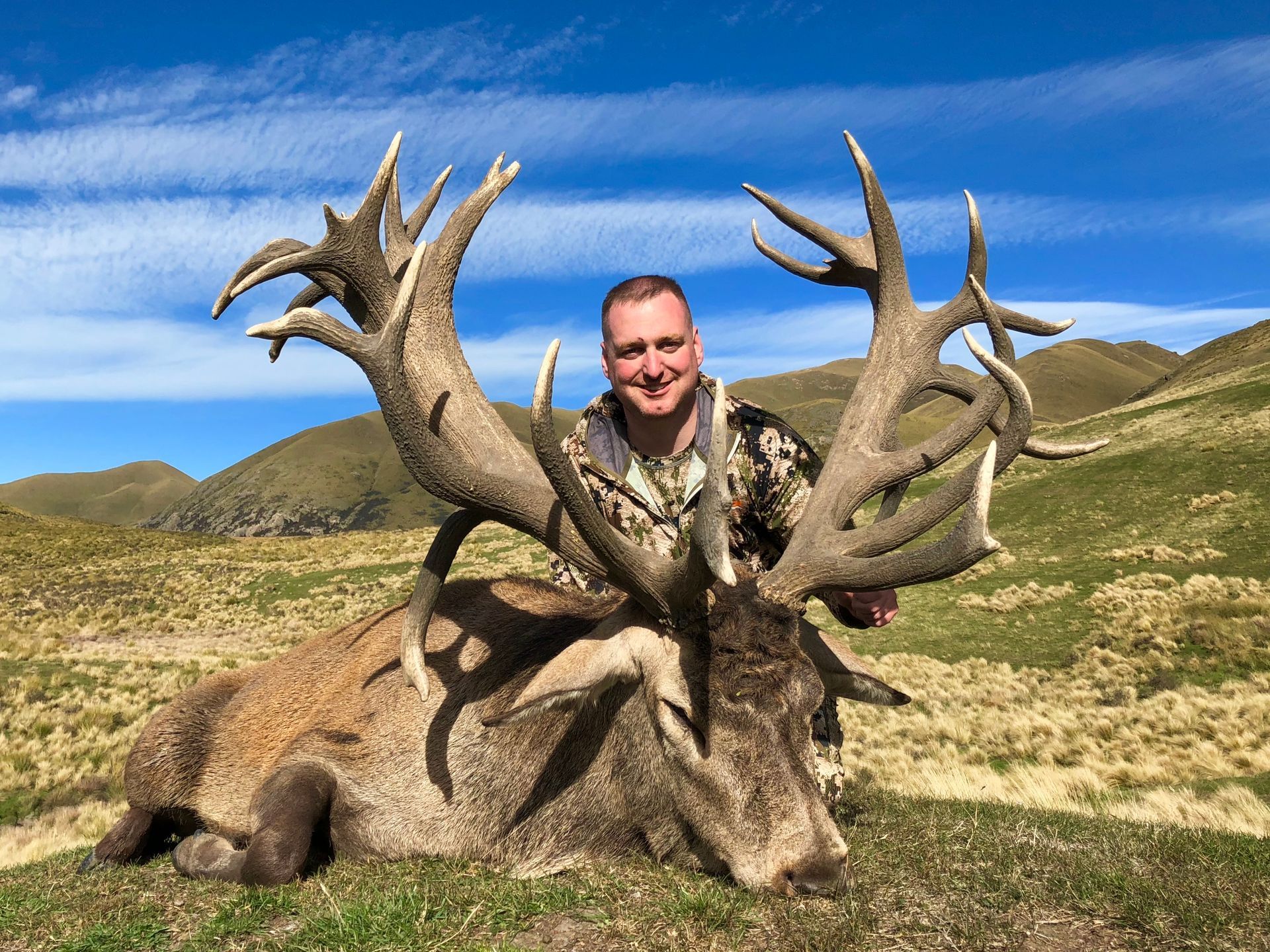 Man smiles, posing with a large red deer he hunted, antlers spanning wide. Hills and blue sky in the background.