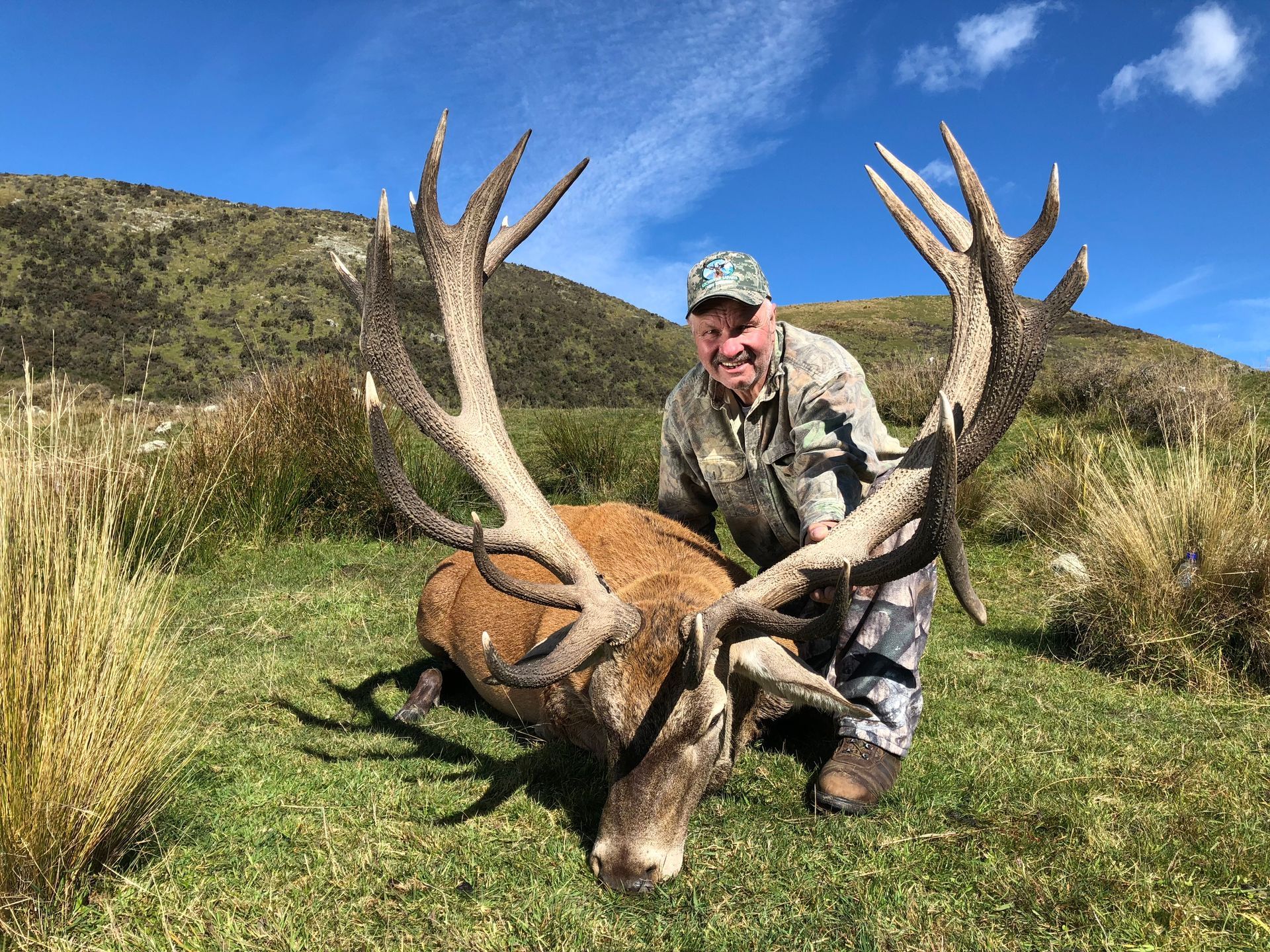 Man in camouflage kneels beside a large red stag with impressive antlers on a grassy hillside, smiling at the camera.