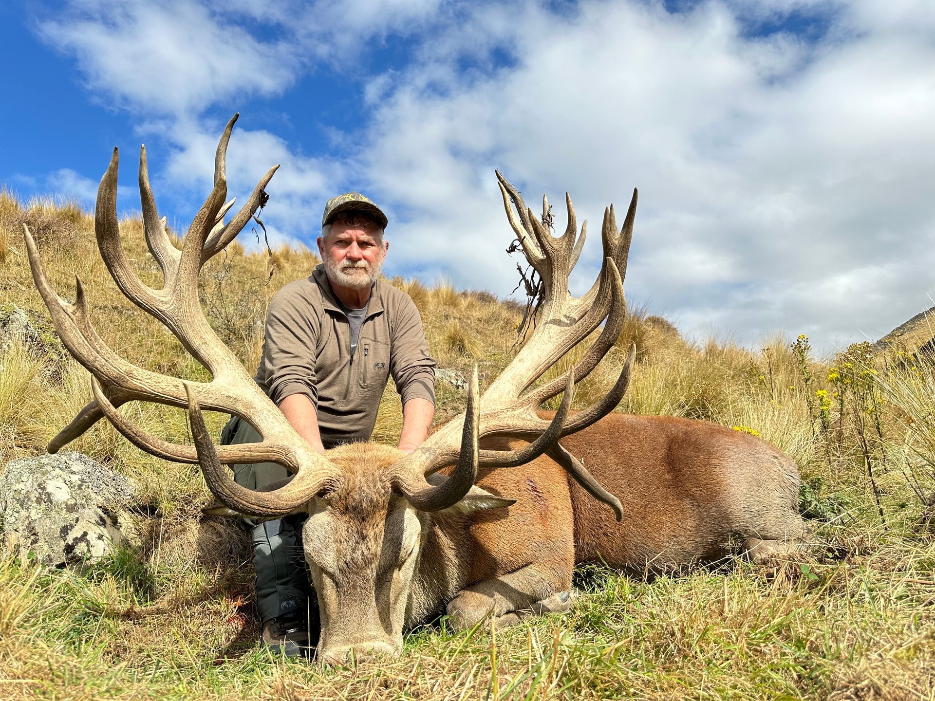Man poses with a large dead red deer, its antlers sprawling.  Grassy hillside setting under a partly cloudy sky.