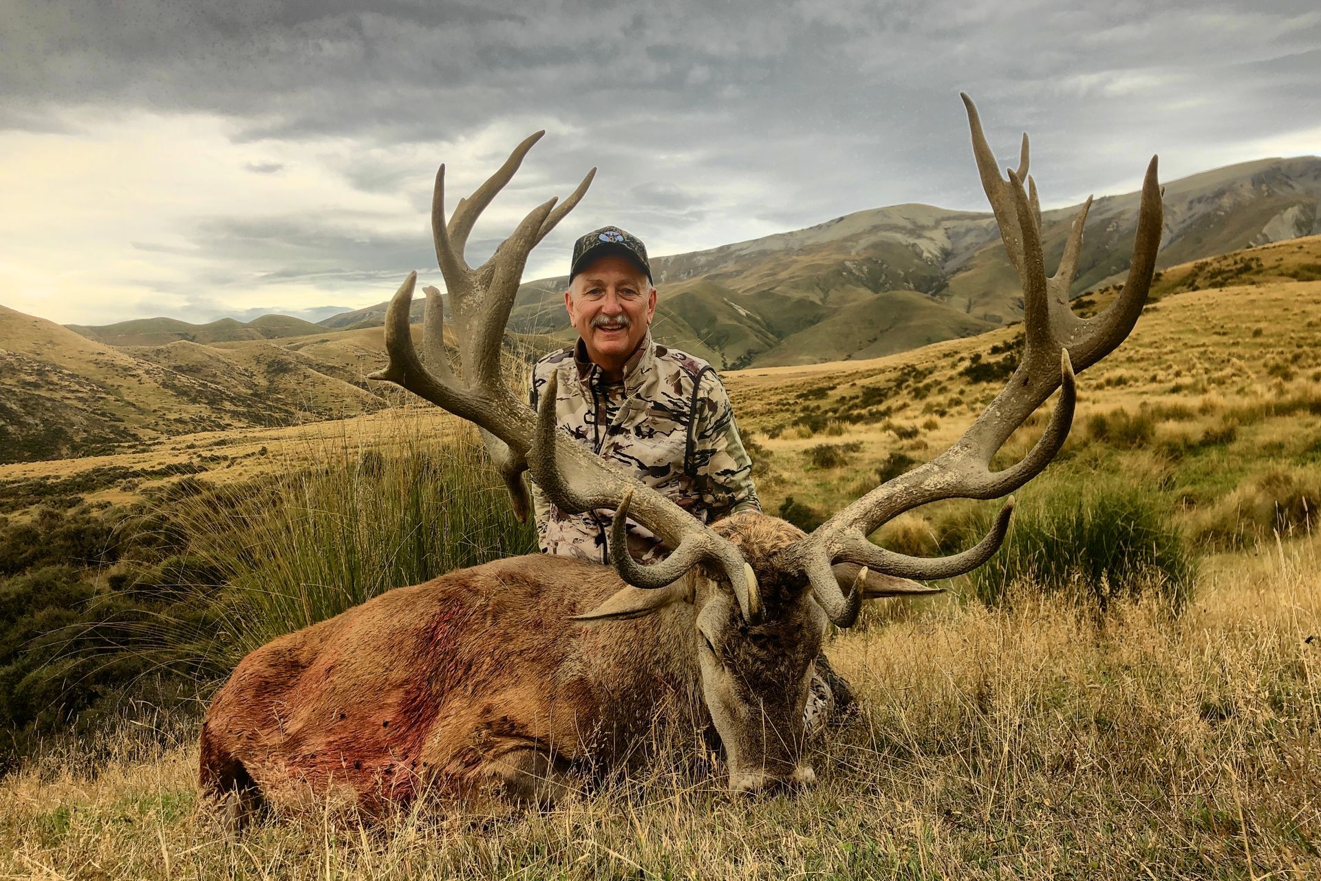 Man in camouflage poses with a large dead red deer in a grassy field, mountain backdrop. Overcast sky.