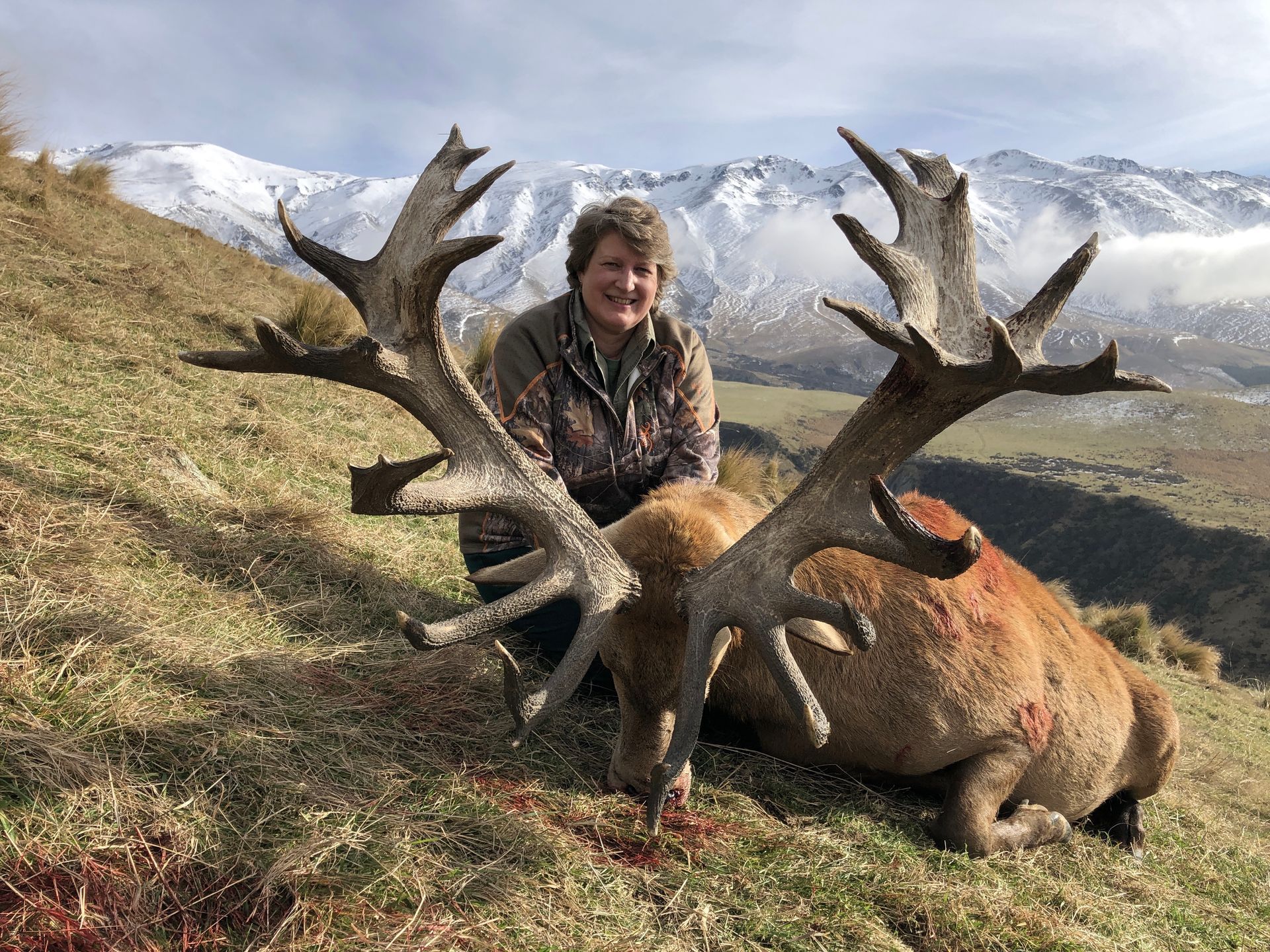 Woman in camouflage kneeling beside a large, harvested stag with impressive antlers, mountains in the background.