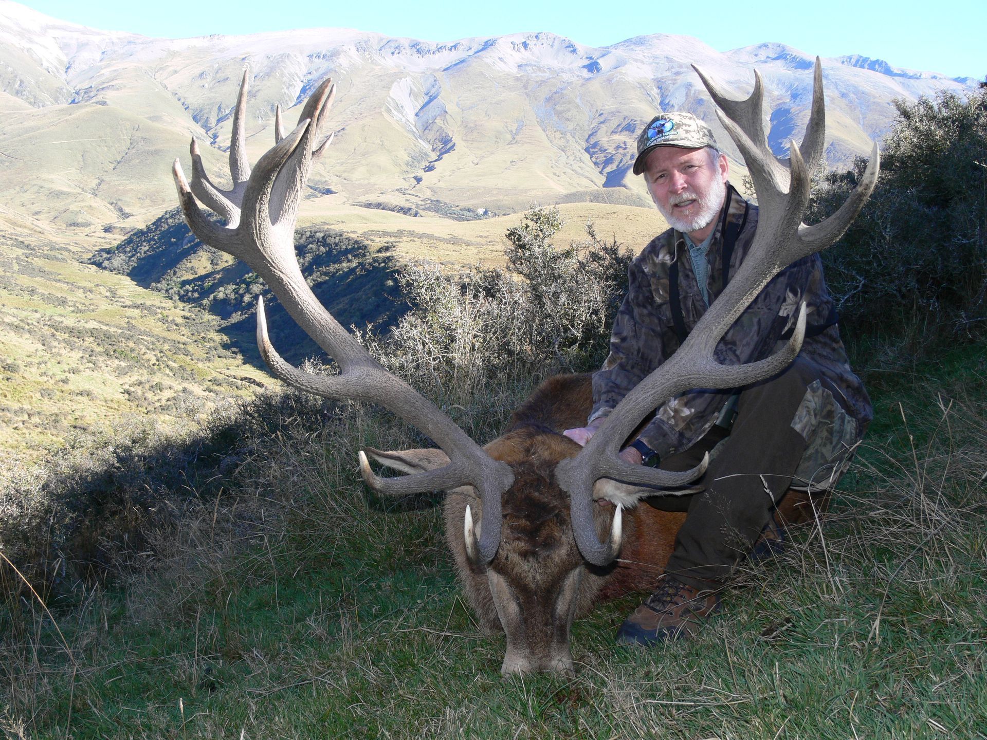 Man kneels beside a large red deer with impressive antlers, posing on a grassy hillside against a mountain backdrop.