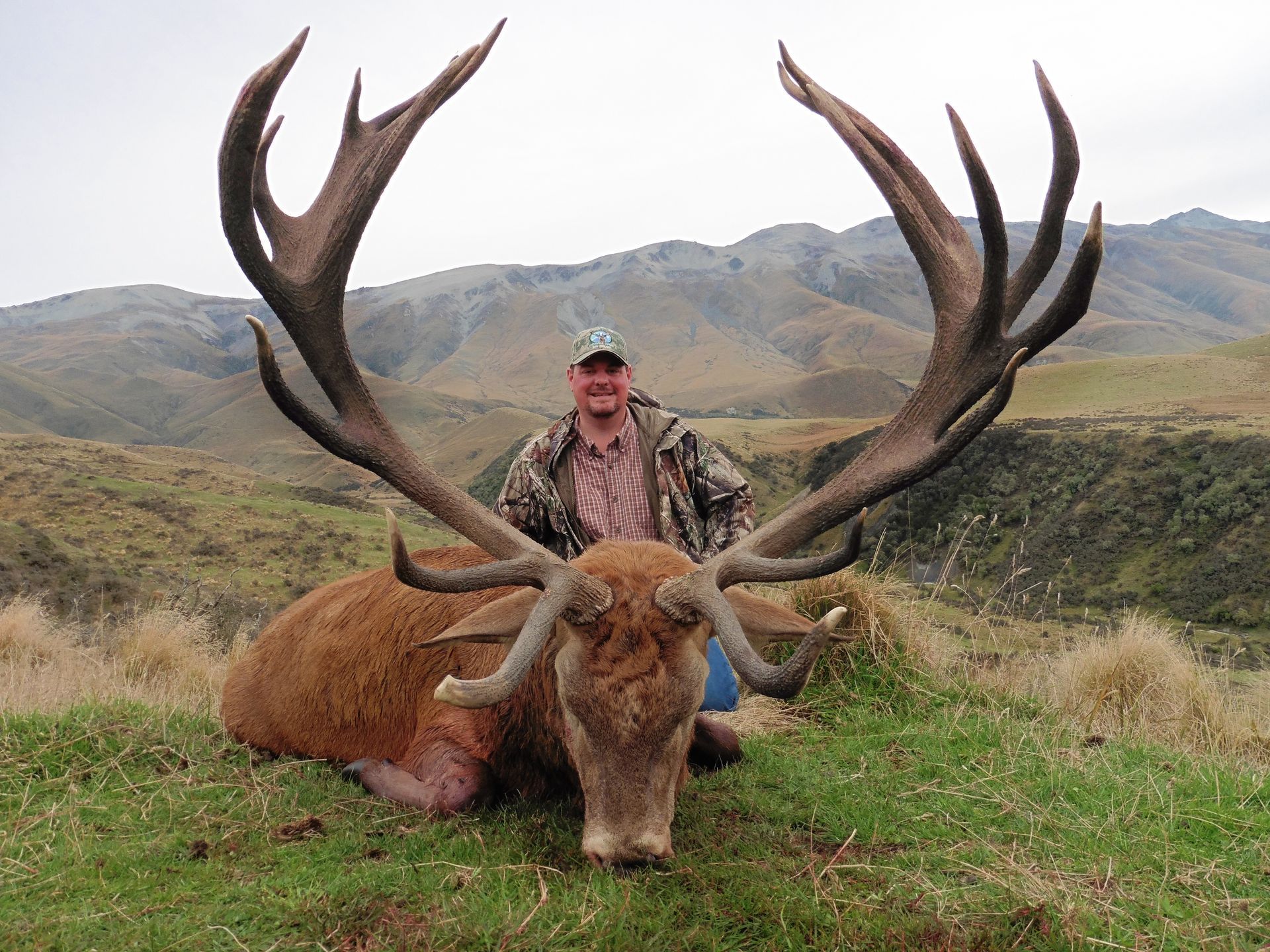 Man poses with a large, dead red deer buck with massive antlers in a grassy, mountainous area. The man is smiling and wearing camouflage.
