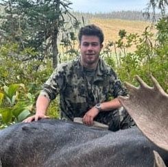 A young man in camouflage sits with a moose, both outdoors. The man smiles, appearing to be hunting.