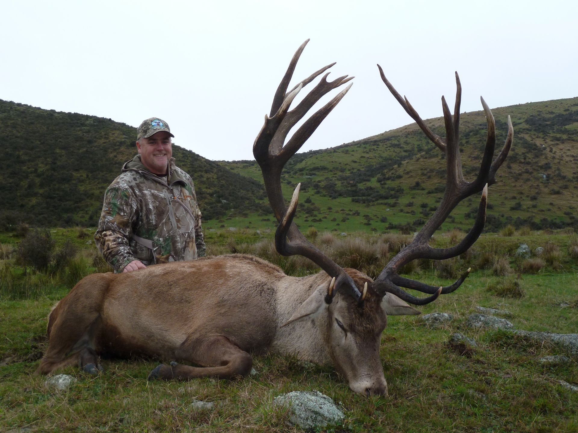Man in camouflage stands next to a large red deer with impressive antlers, lying on green grass, in a hilly, outdoor setting.