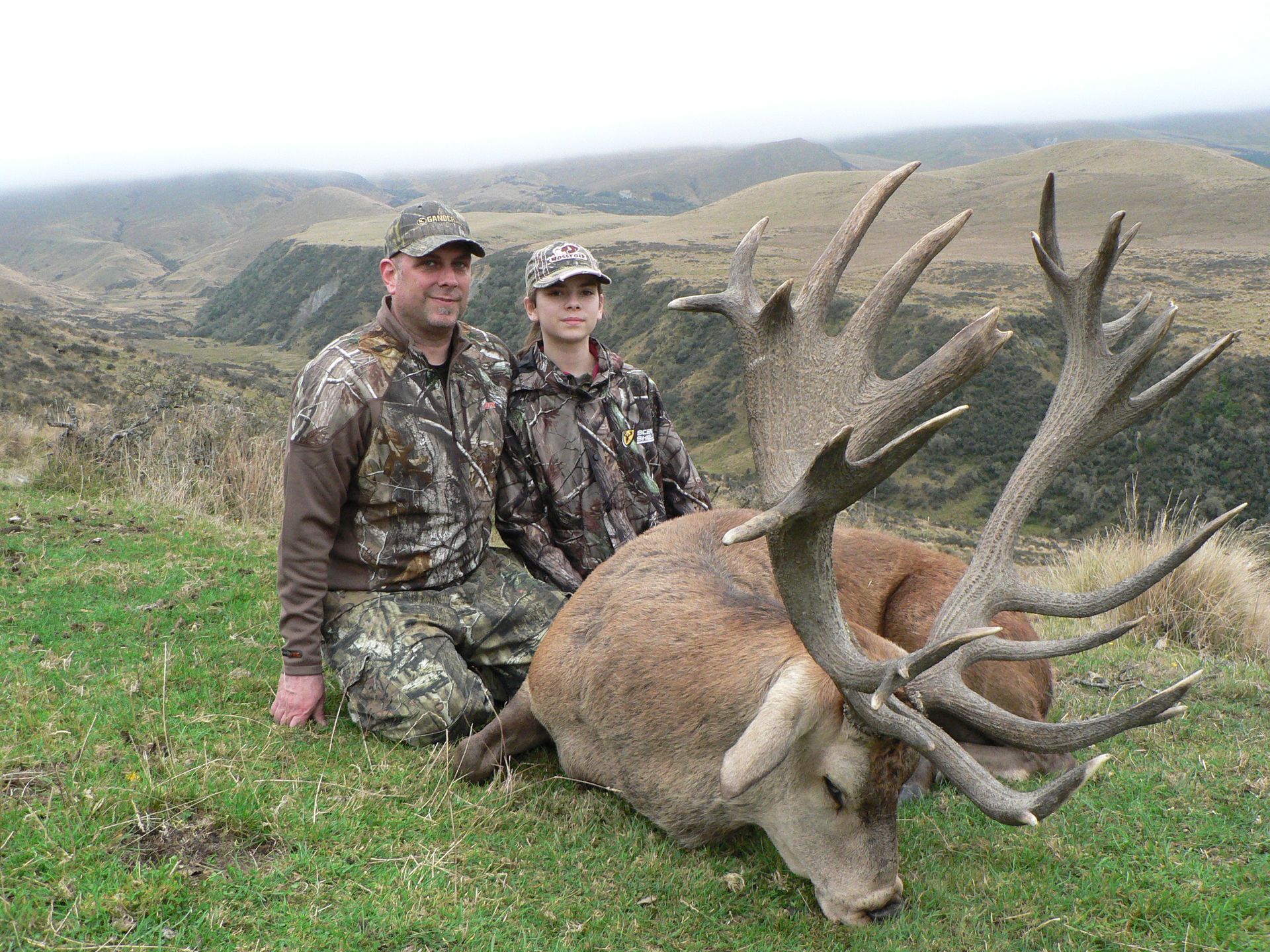A man and a youth in camouflage pose with a large red deer stag on a grassy hillside, with a valley in the background.