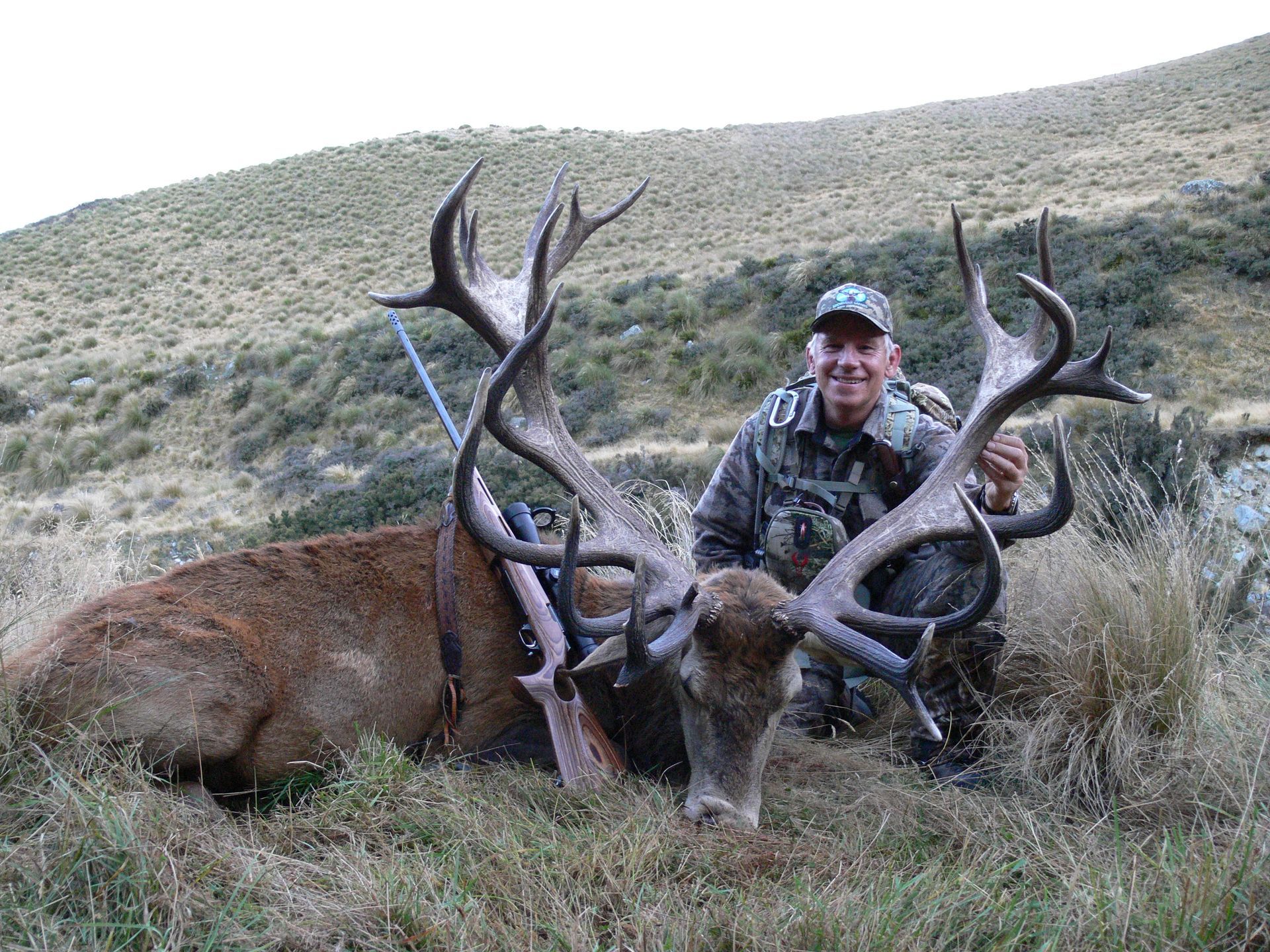 A man in camouflage kneels beside a large red deer with impressive antlers in a grassy mountain setting, smiling.