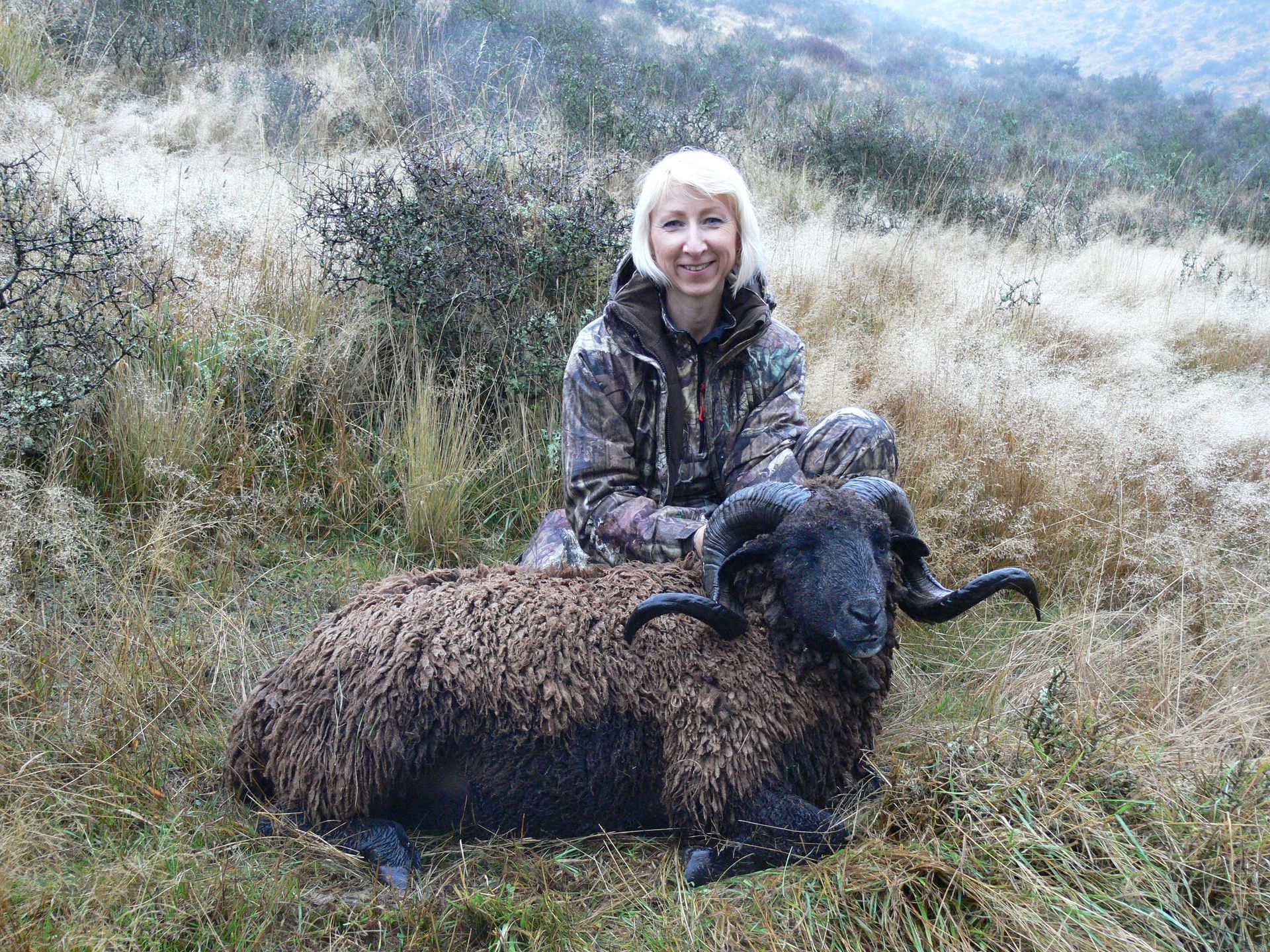 Woman in camouflage sits beside a dark-wooled ram in a grassy field. The woman smiles.