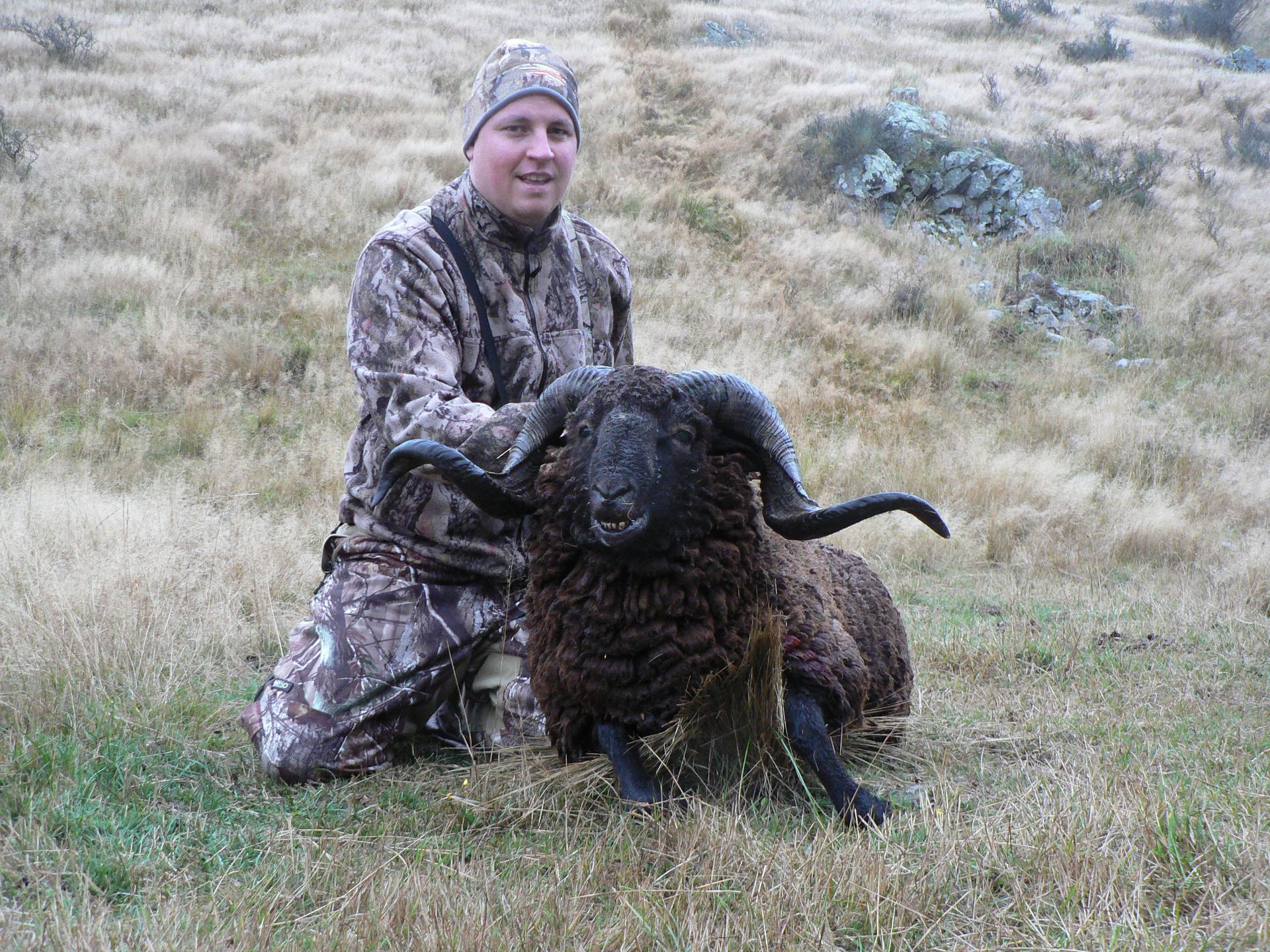 A man in camouflage kneels beside a black ram with large horns, smiling on a grassy hillside.