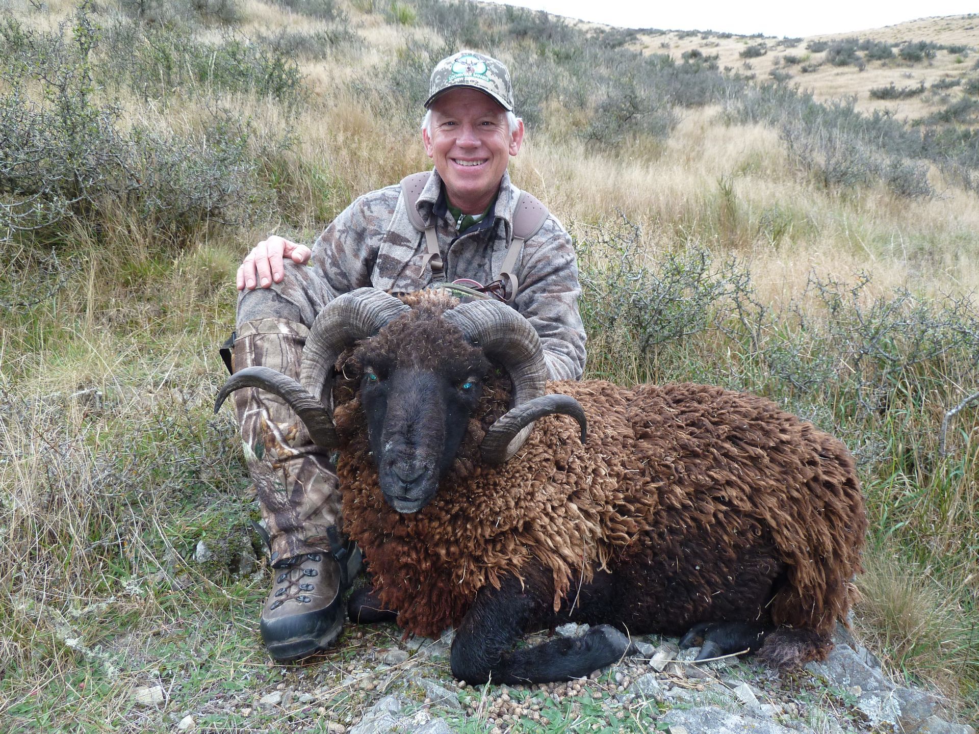 Man in camouflage kneeling beside a dark-haired ram with large curled horns on a grassy hillside. He is smiling.