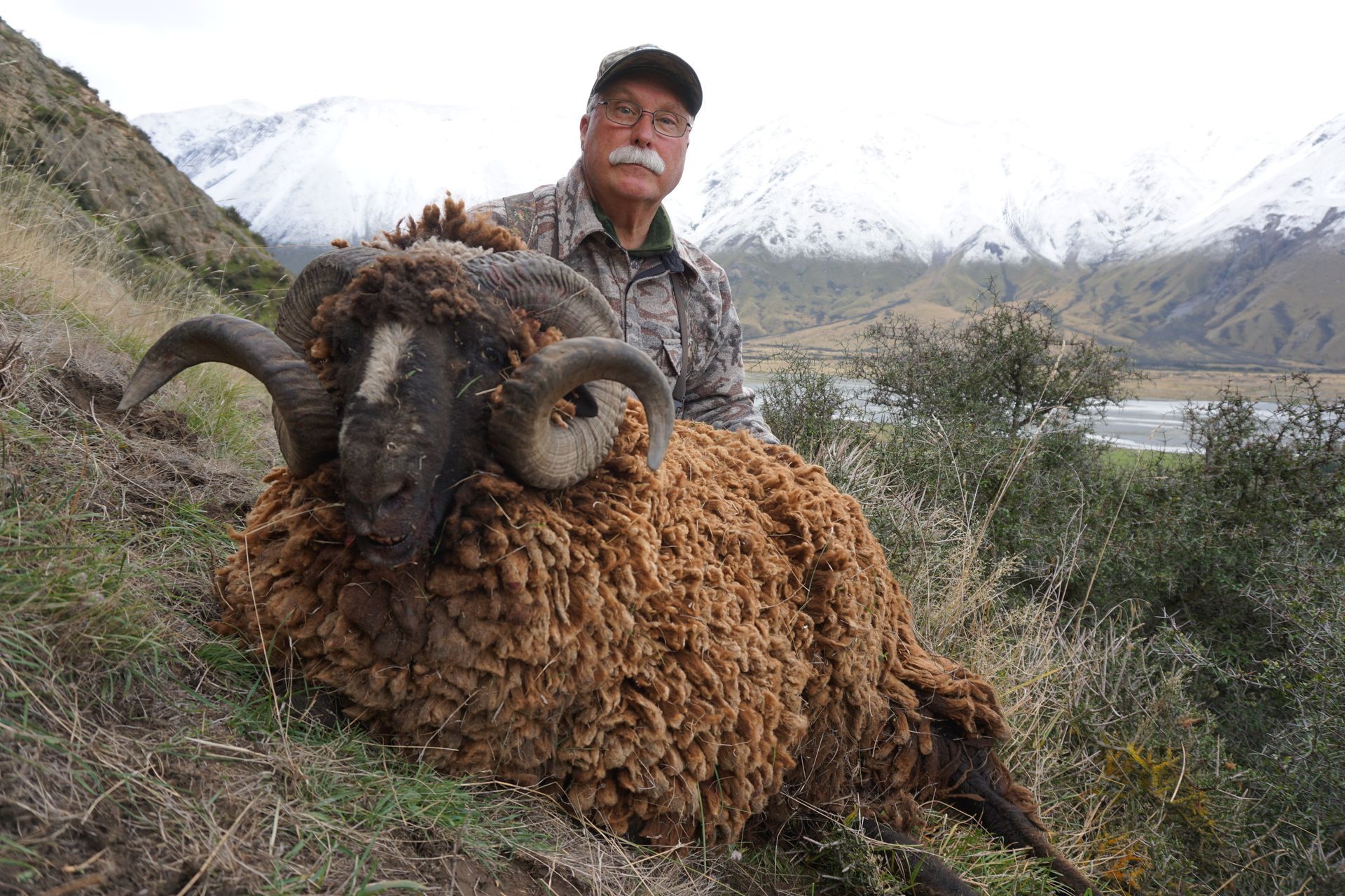 Man with mustache in camouflage holds a large, brown-fleeced ram with large horns, in a mountainous, snowy landscape.